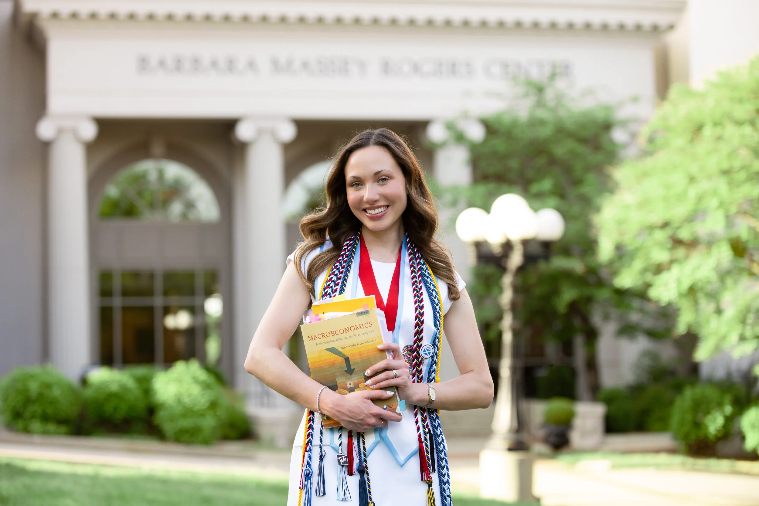 A young woman in a white dress standing outside in front of the Barbara McClintock Roche Center, smiling, holding books and wearing medallions and other graduation cords.