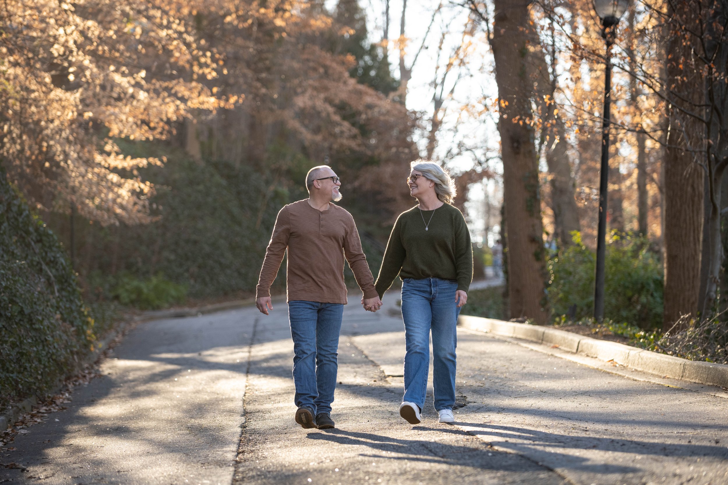 A couple holding hands and walking on a paved path through a park with trees and fall foliage, smiling at each other in the late afternoon sunlight.