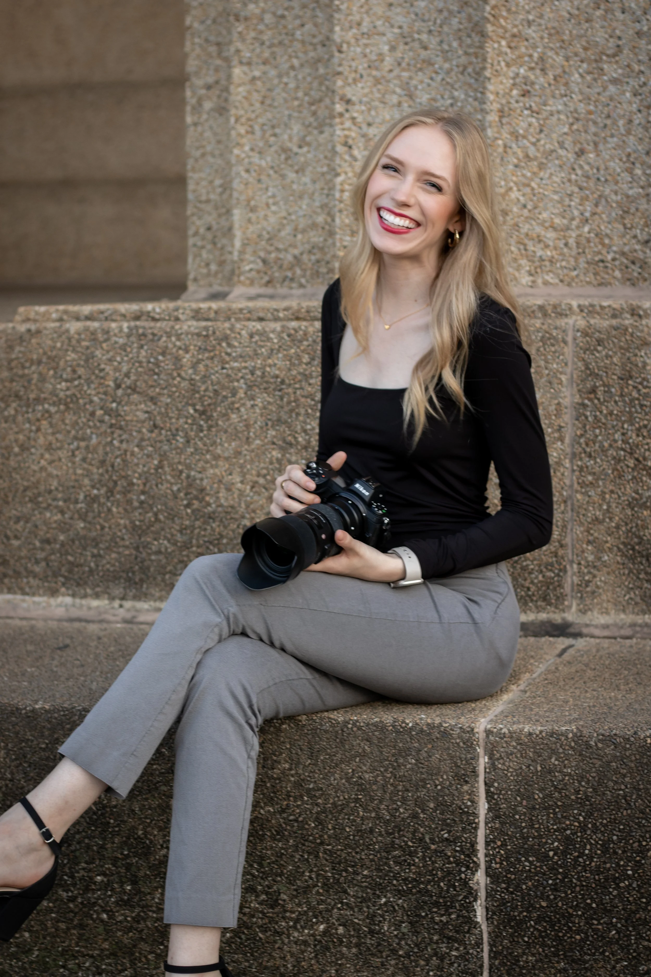 A smiling young woman sitting on a stone ledge, holding a professional camera with a large lens, wearing a black long-sleeve top, gray pants, and black shoes.