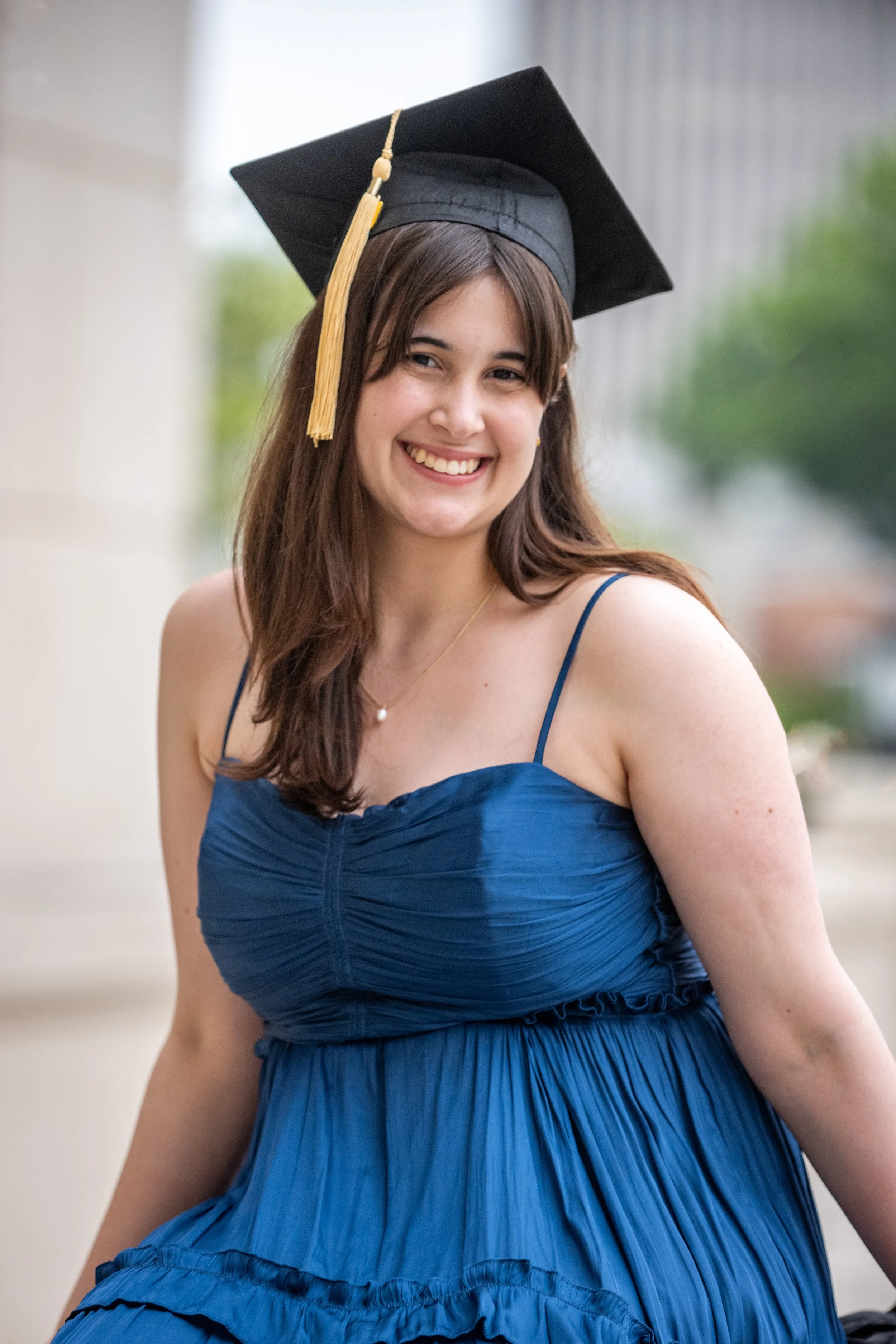 Young woman in a blue dress wearing a black graduation cap with a gold tassel, smiling outdoors.