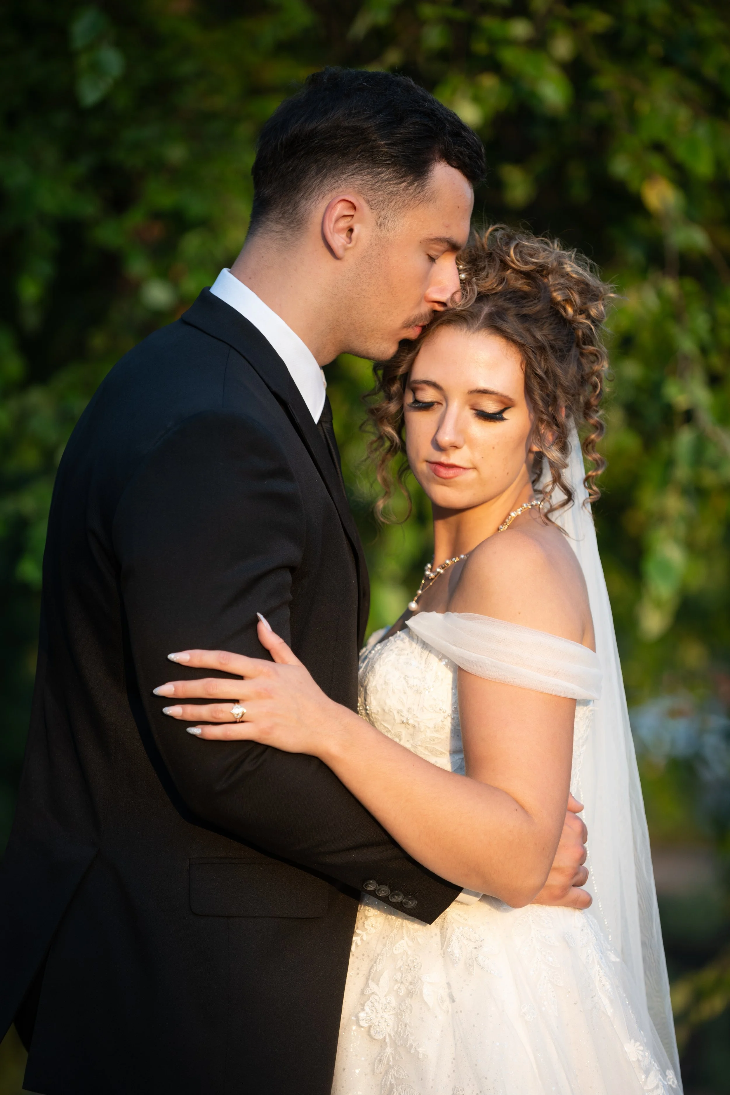 A bride and groom embrace outdoors with their eyes closed, standing close together, with green foliage in the background.