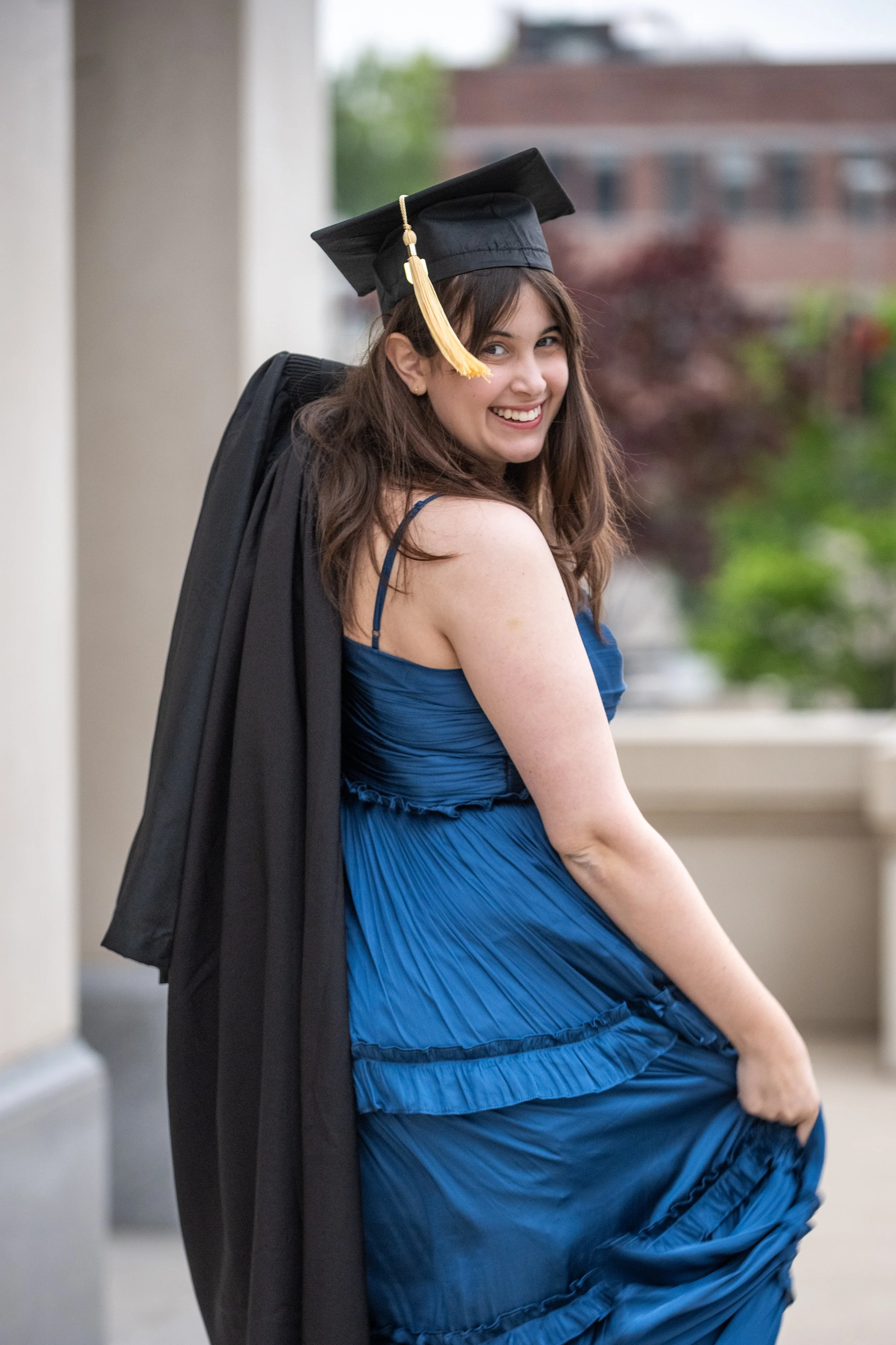 A young woman in a blue dress and graduation cap, smiling, with a black academic gown draped over her shoulder.