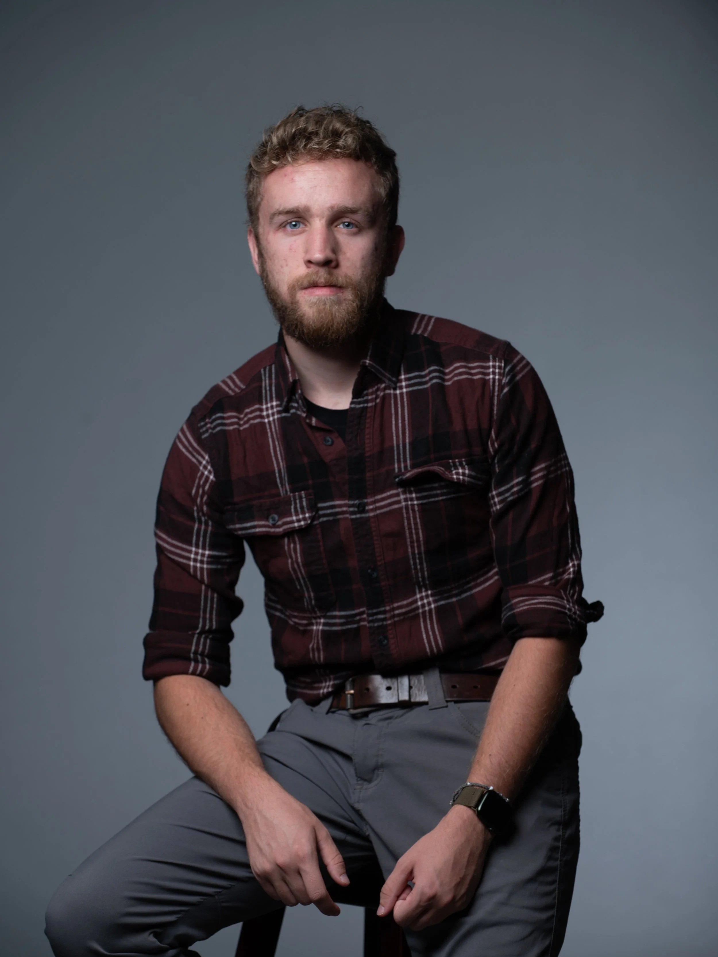 A young man with curly blonde hair and a beard, wearing a red plaid shirt with rolled-up sleeves and gray pants, sitting on a stool against a plain gray background.