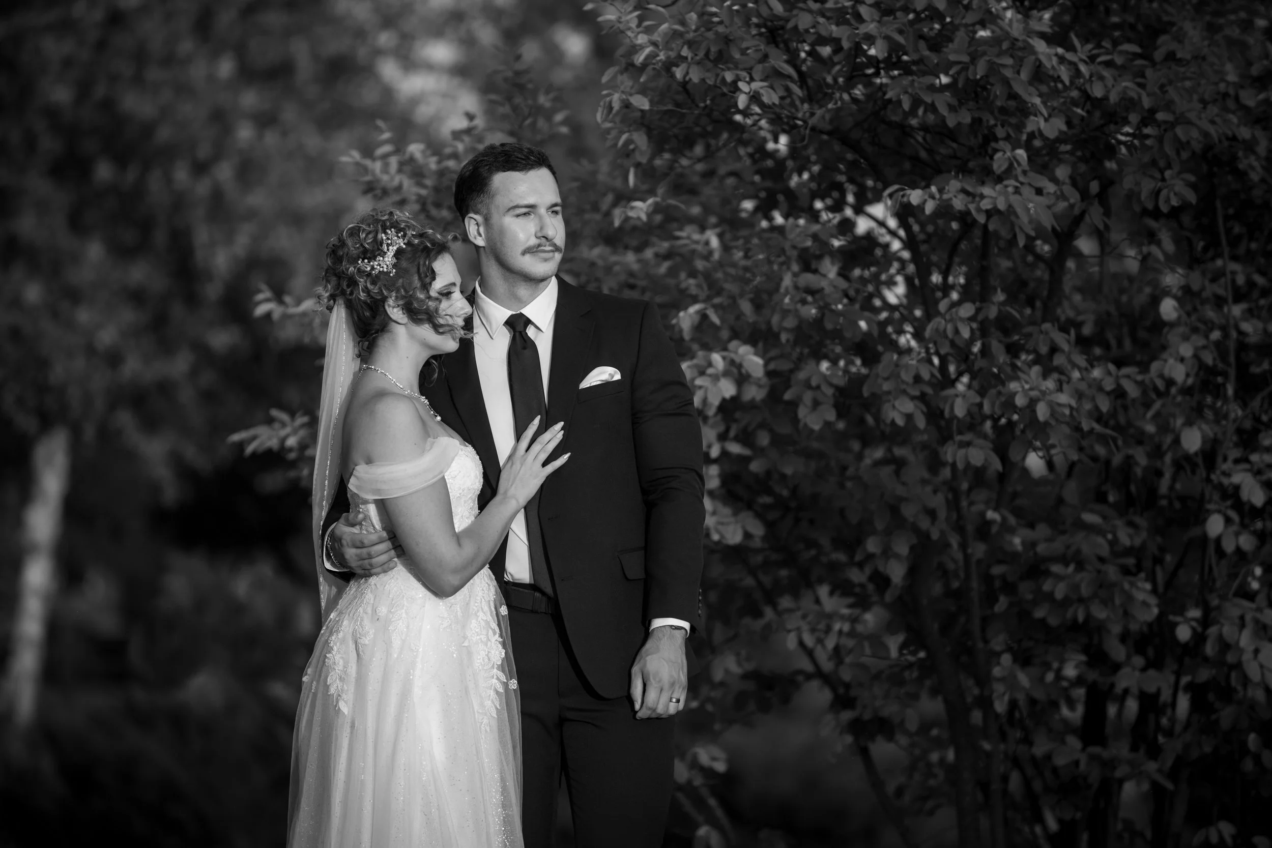 A black and white photograph of a bride and groom standing outdoors, with the bride leaning her head on the groom's shoulder and both looking into the distance, surrounded by trees and foliage.