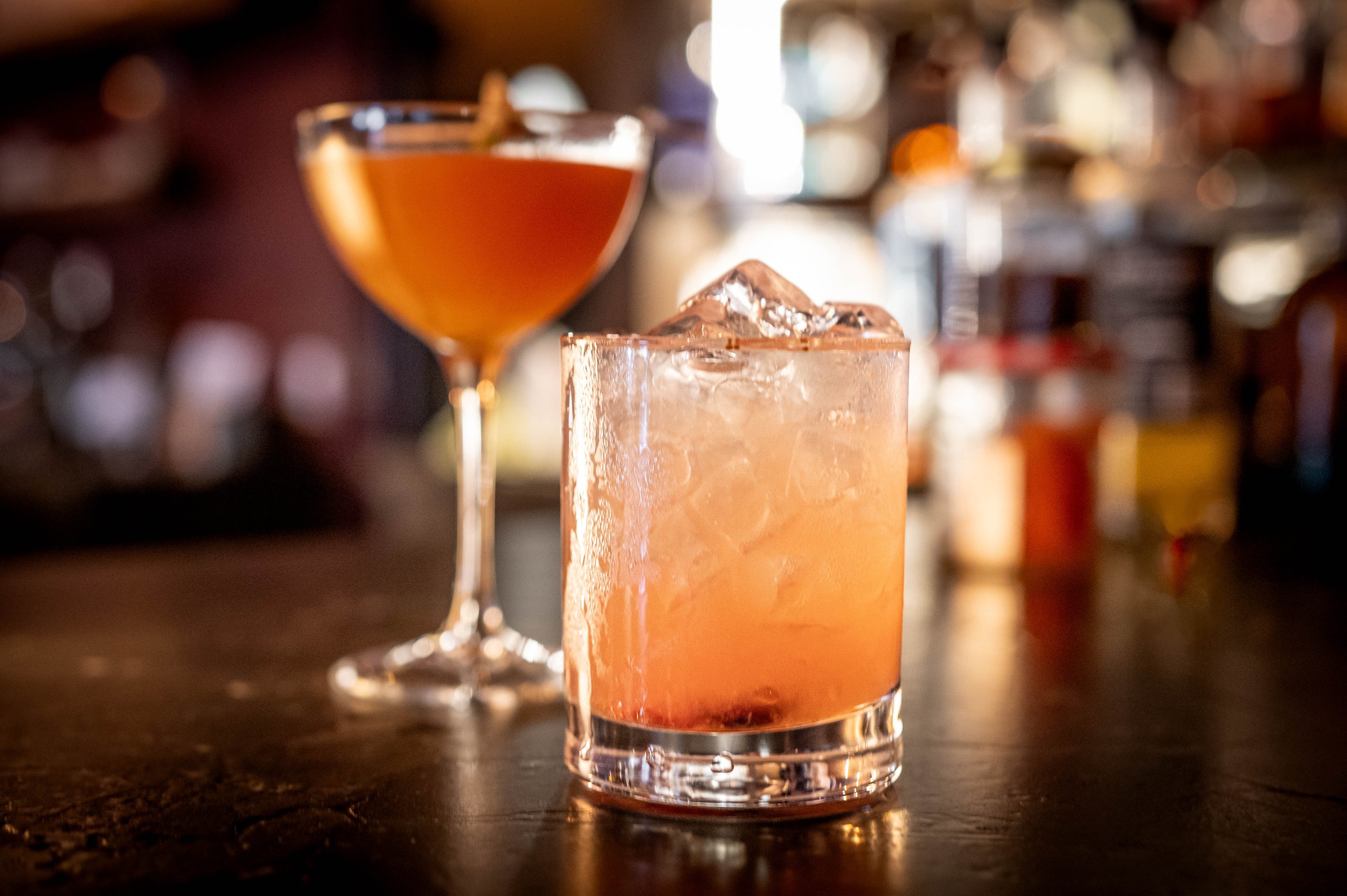 Close-up of a cocktail in a glass with ice, in front of a second cocktail on a dark bar counter with a blurred background of bar equipment and bottles.