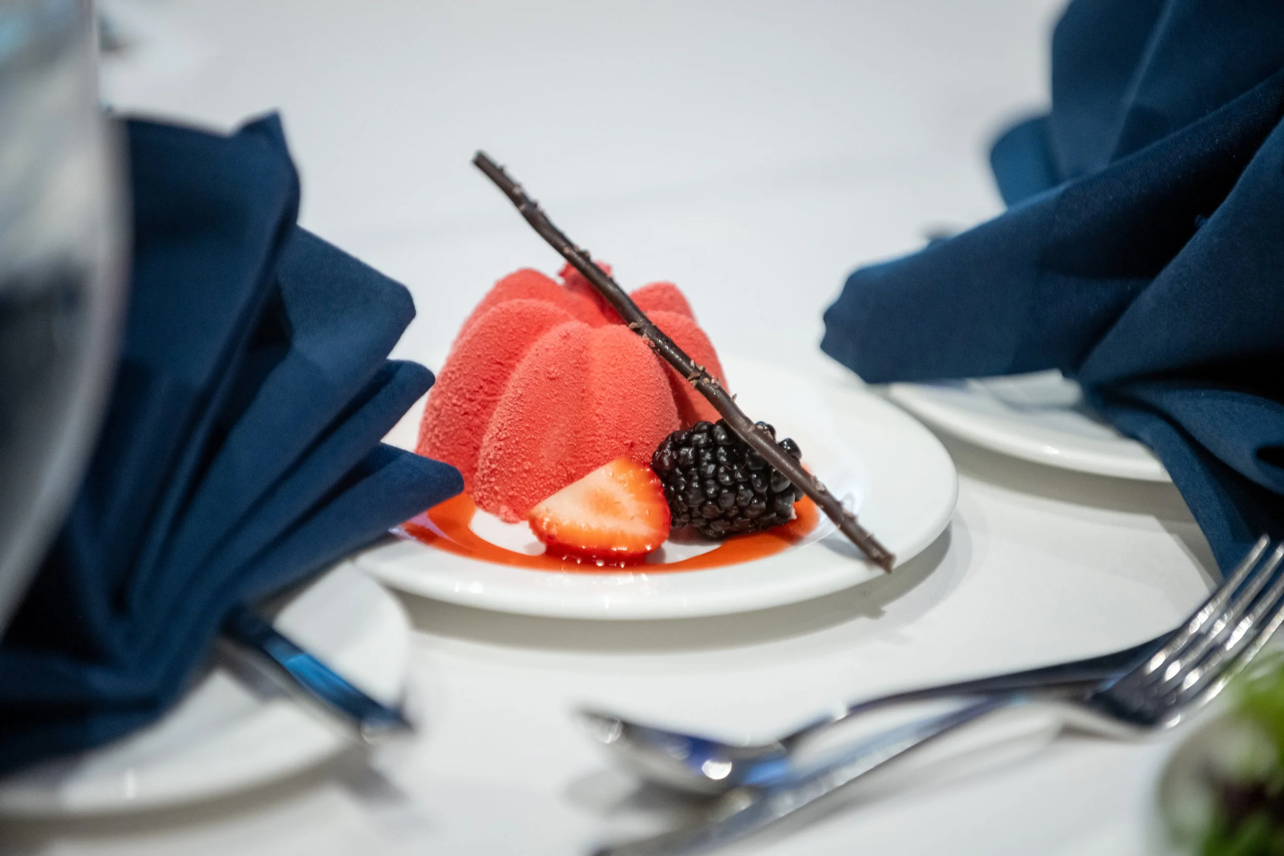 A plated dessert with red mousse, strawberries, blackberries, and a decorative chocolate stick on a white plate, surrounded by navy blue napkins and serving utensils.