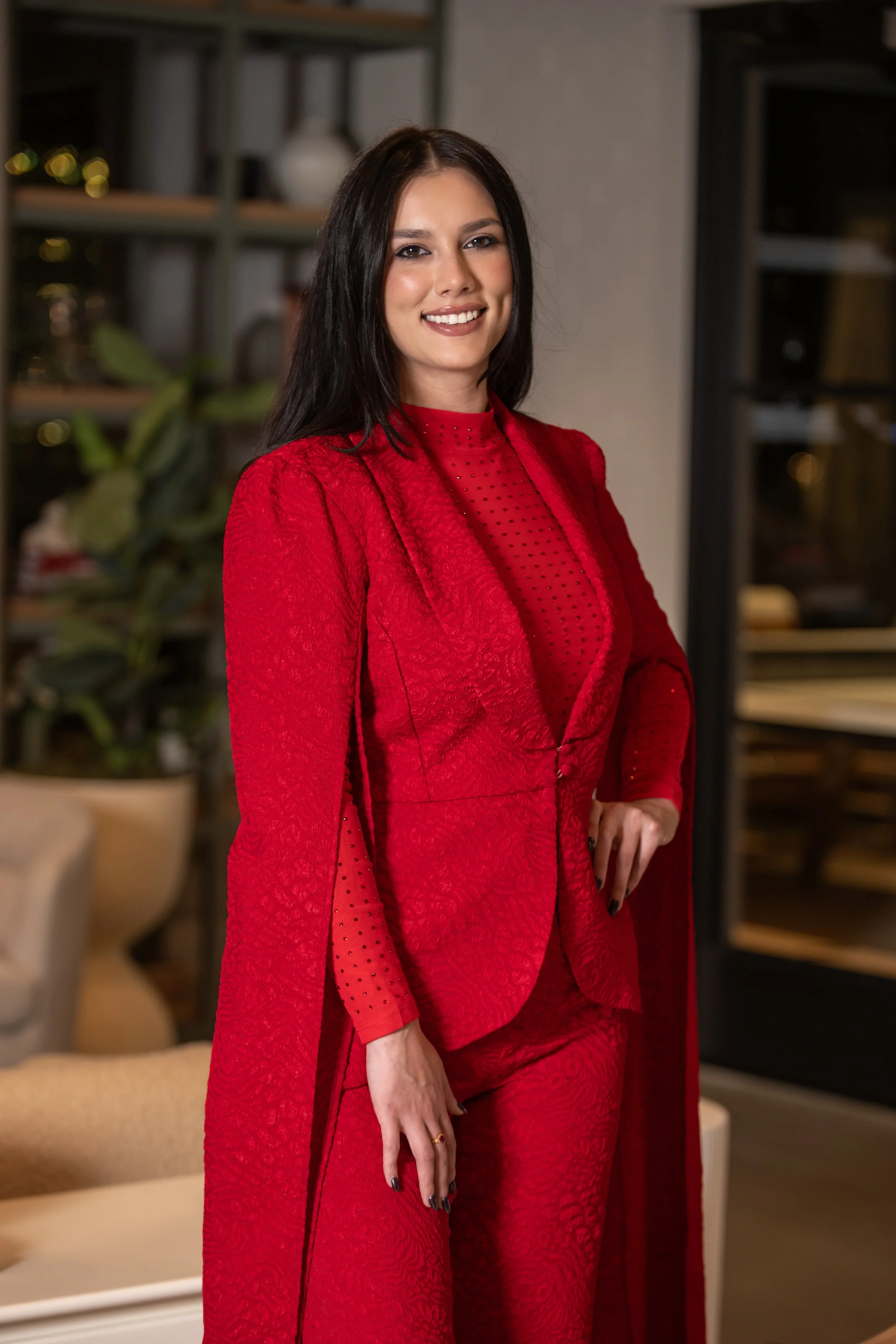 A woman in a red suit and matching pants, with a sheer polka dot top underneath, standing indoors with a confident smile.