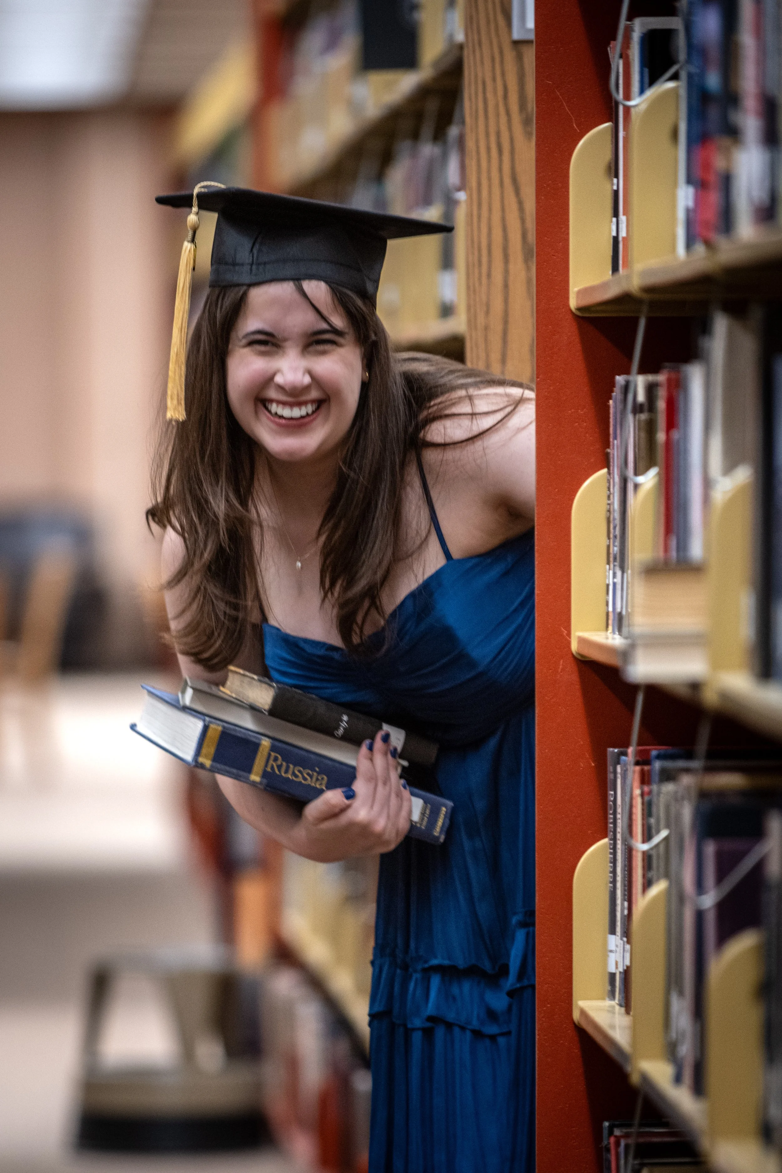 A young woman in a blue dress and graduation cap peeks around a bookshelf in a library, smiling while holding books.