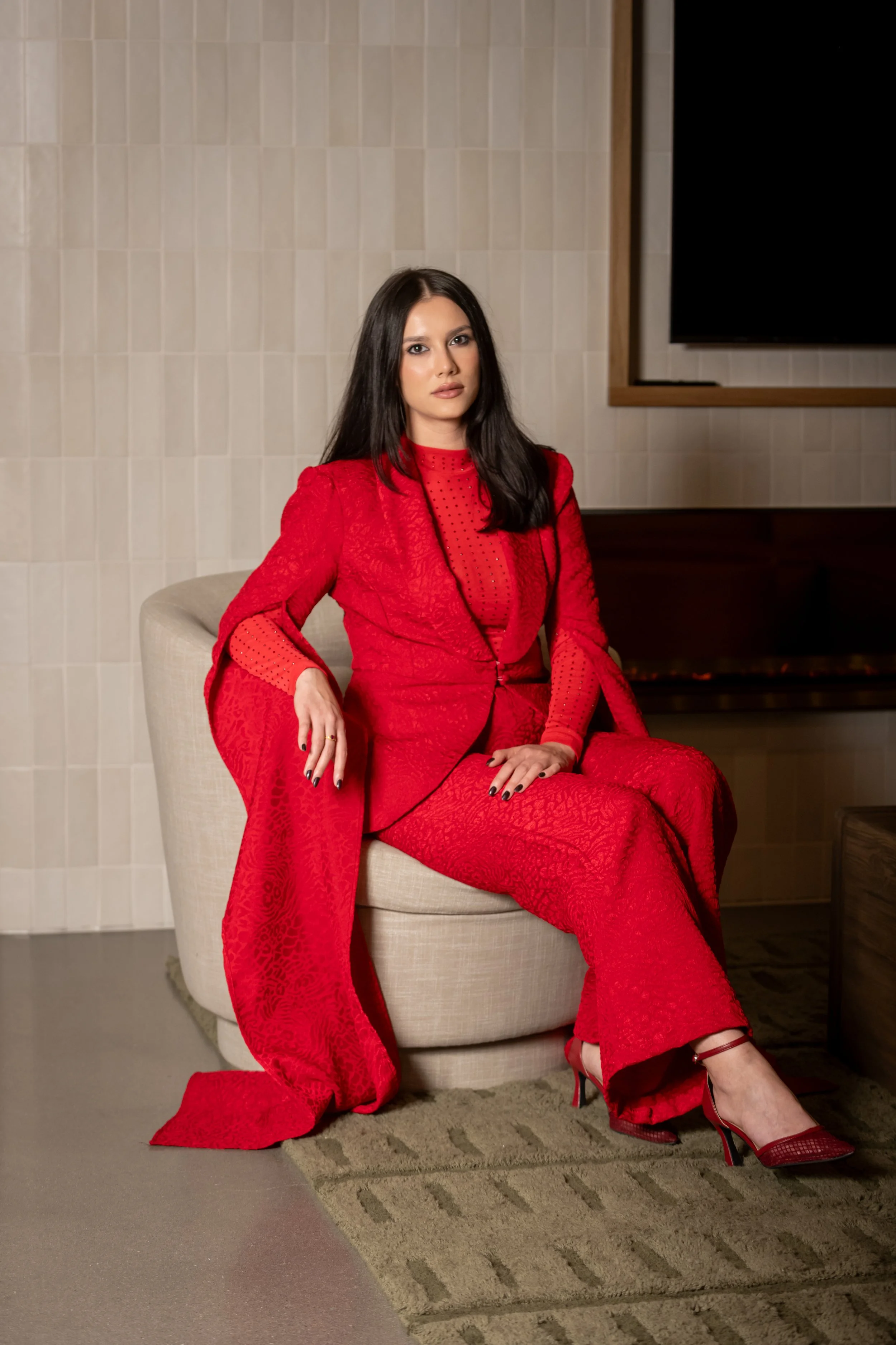 A woman with long dark hair wearing a red textured suit and red high heels, seated on a beige armchair in a room with a light-colored tiled wall in the background.