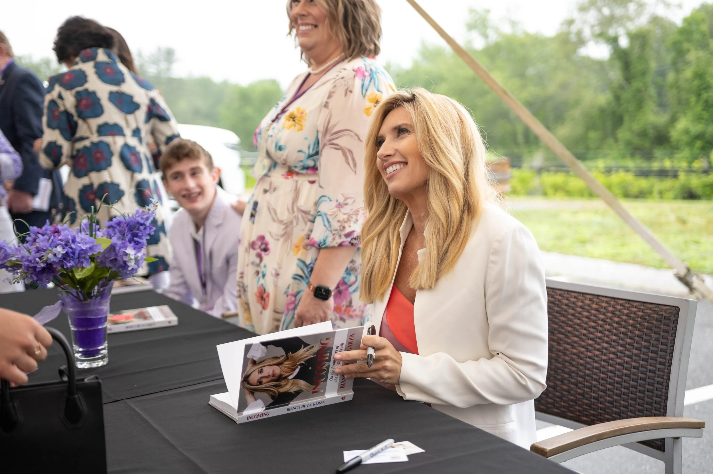 A woman with long blonde hair wearing a white blazer and coral top, smiling while signing books at an outdoor signing event. Other people and a table with purple flowers are visible in the background.