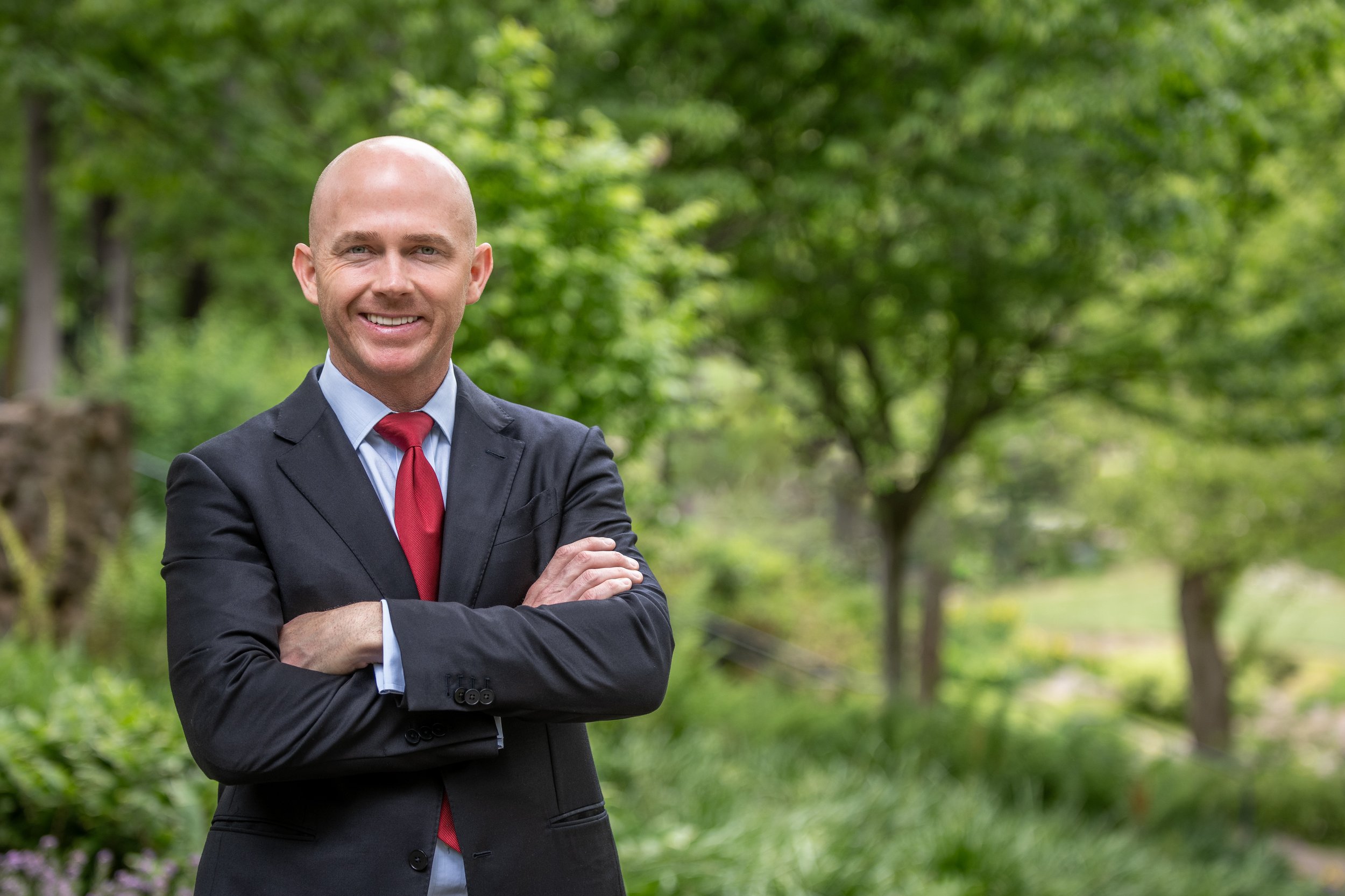 A bald man in a dark suit, light blue shirt, and red tie, standing outdoors with crossed arms and smiling in front of green trees and foliage.
