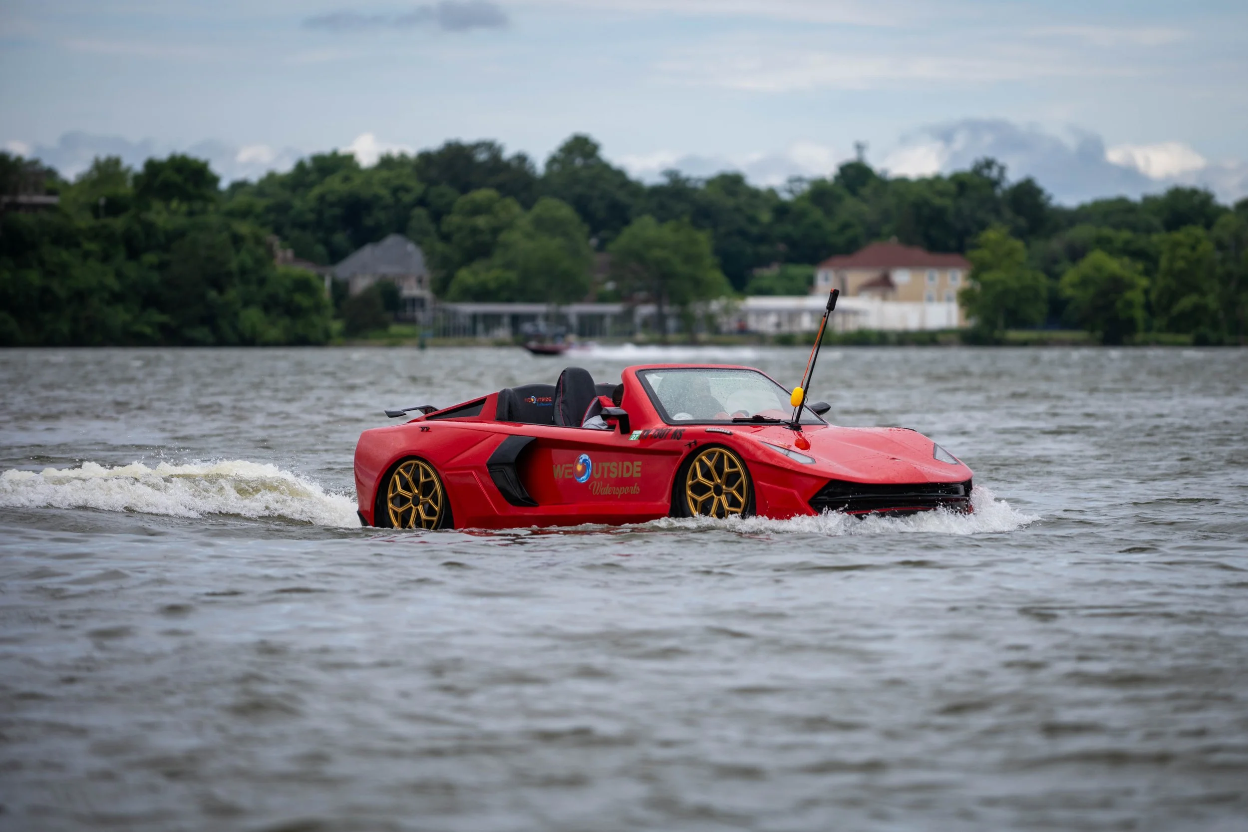 A red sports car designed for water racing is moving across a body of water, creating small waves. The car has gold wheels, a black interior seat, and a long antenna on the front. There are green trees and houses in the background under a cloudy sky.
