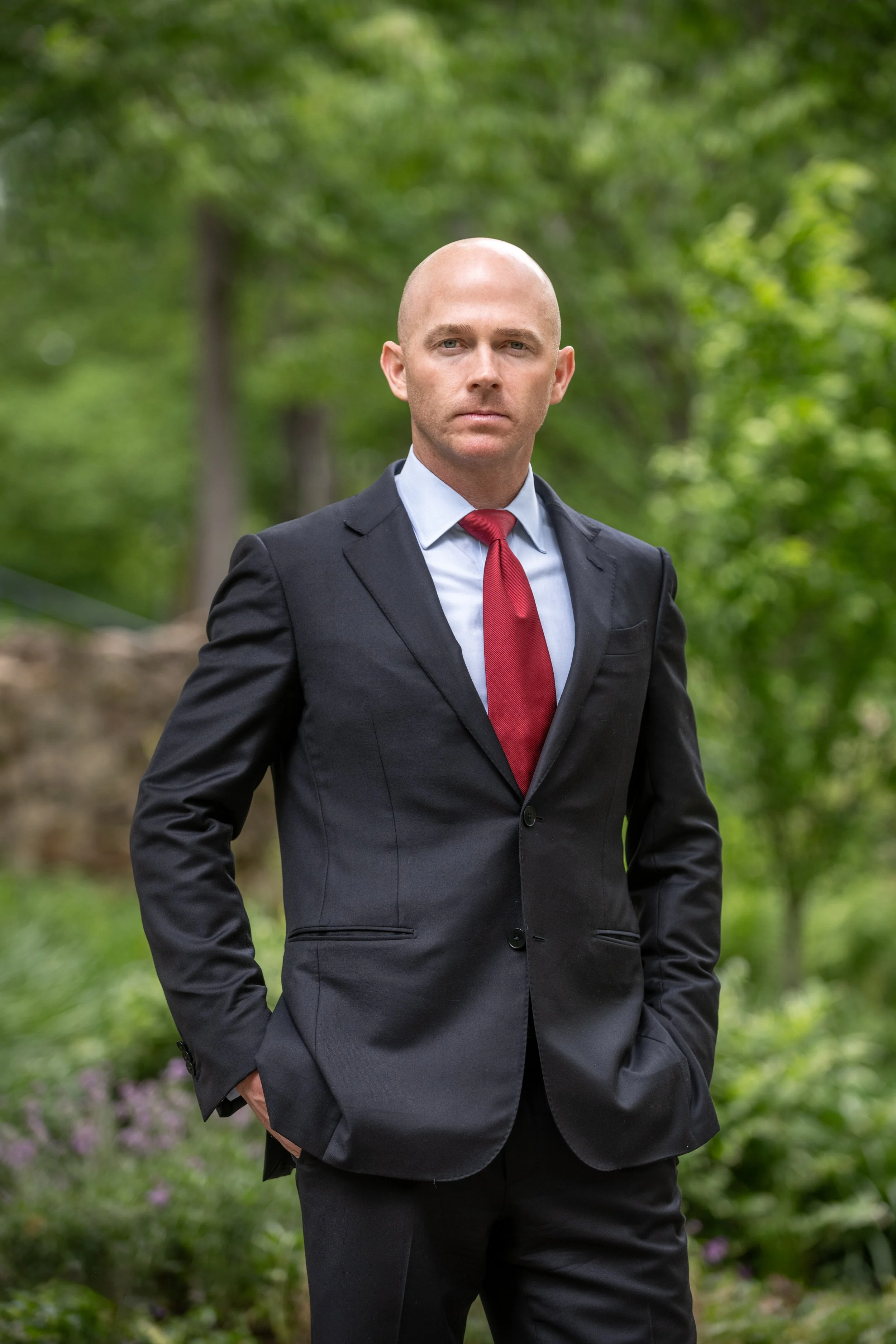 A man in a black suit with a red tie standing outdoors in a green, wooded area.