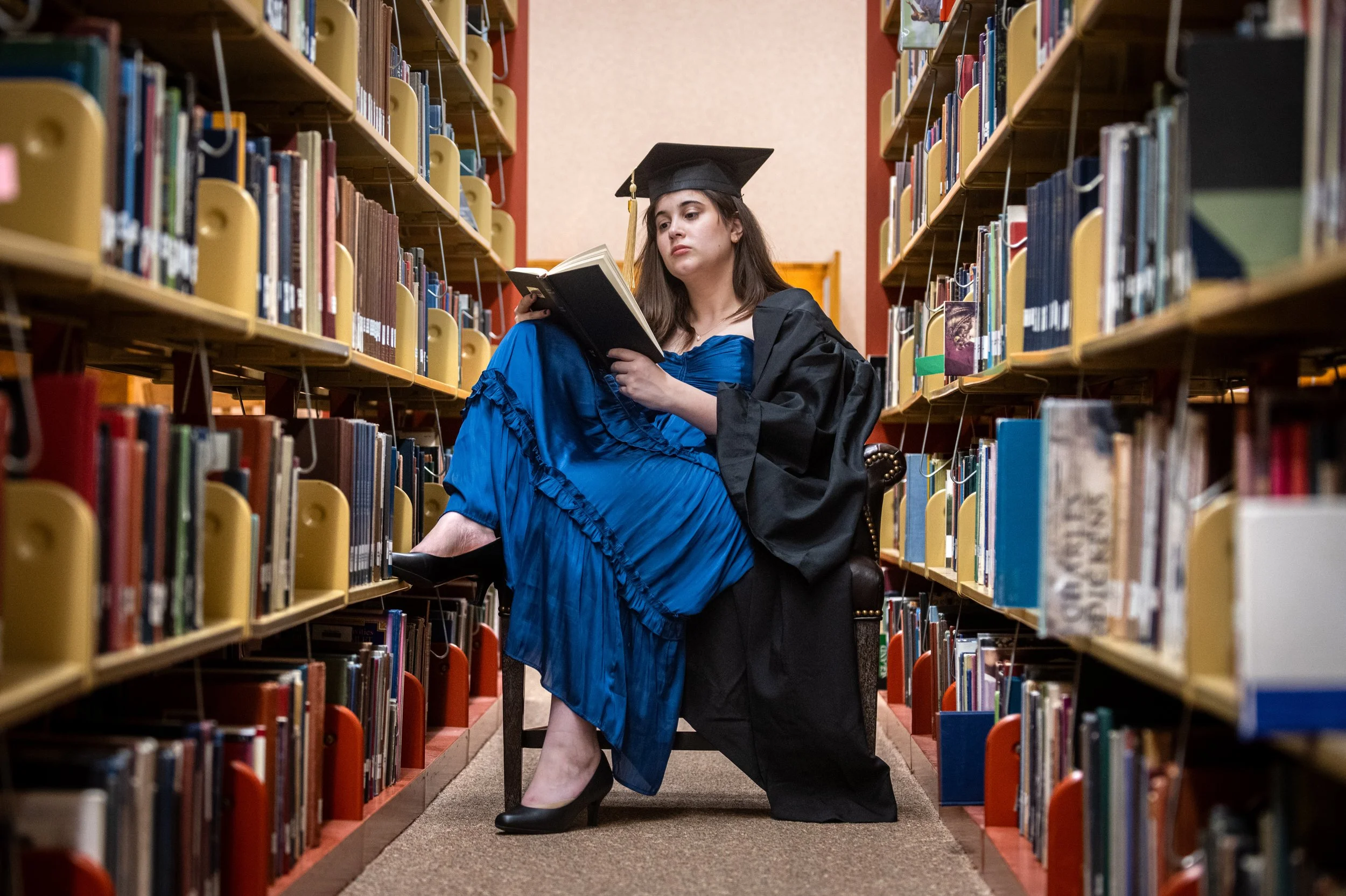 A woman in a graduation gown and cap reading a book while sitting on a chair in a library aisle.