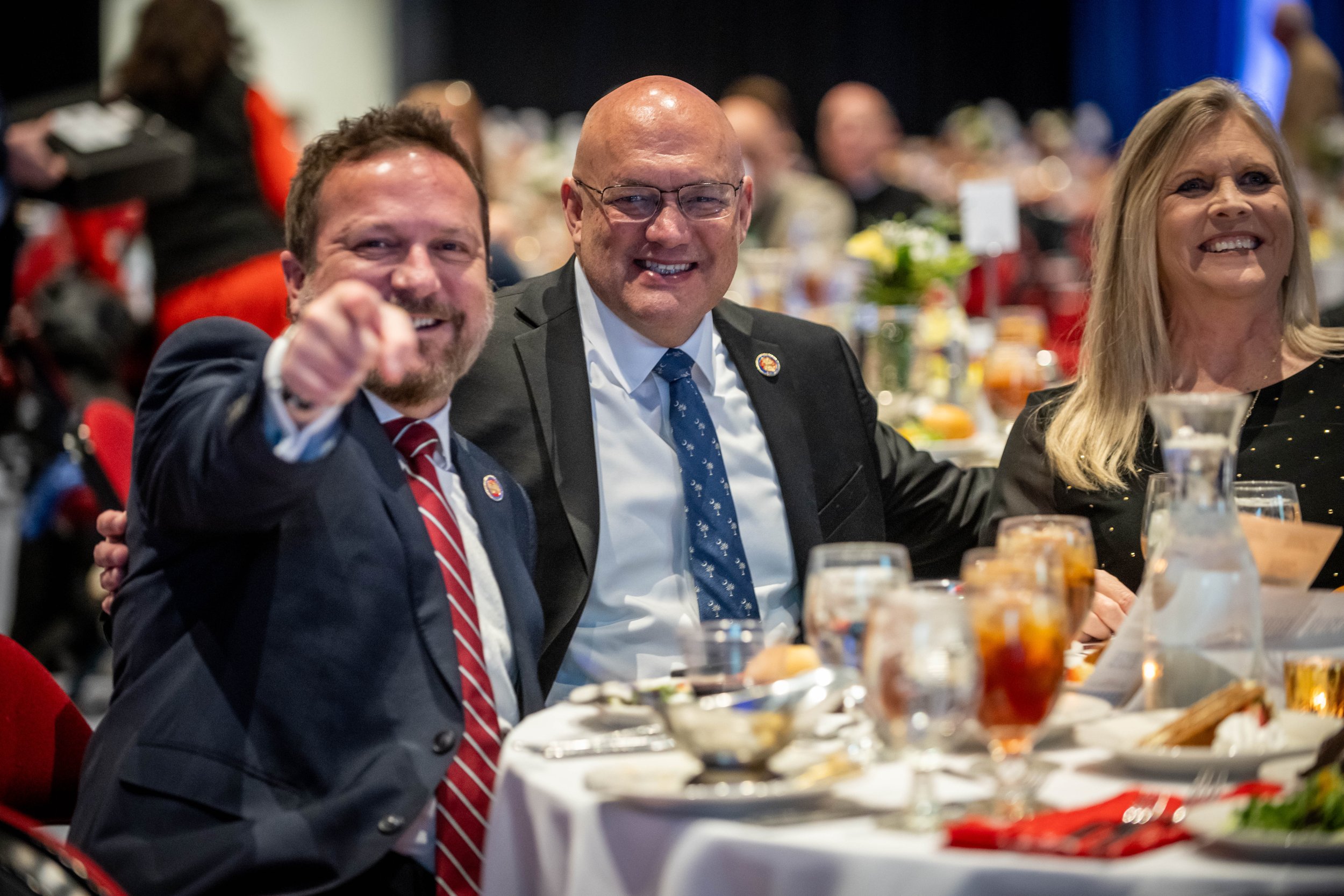Three people sitting at a banquet table, smiling and enjoying a social event, with food and drinks on the table.