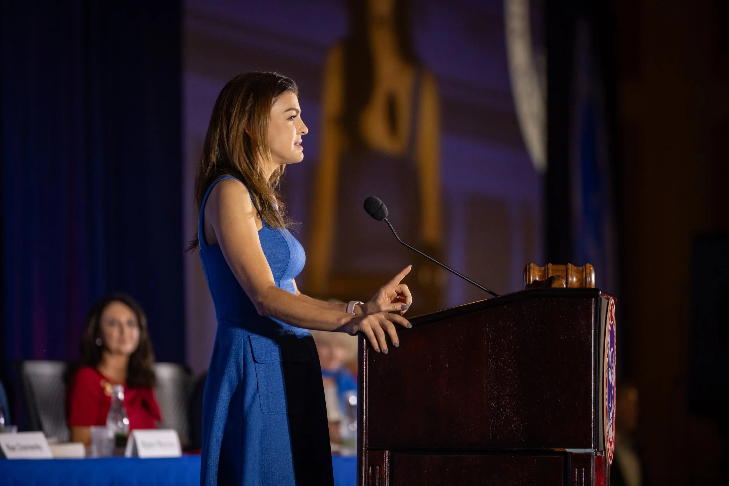 A woman in a blue dress speaking at a podium during a conference or event, with a woman in a red dress seated behind her.