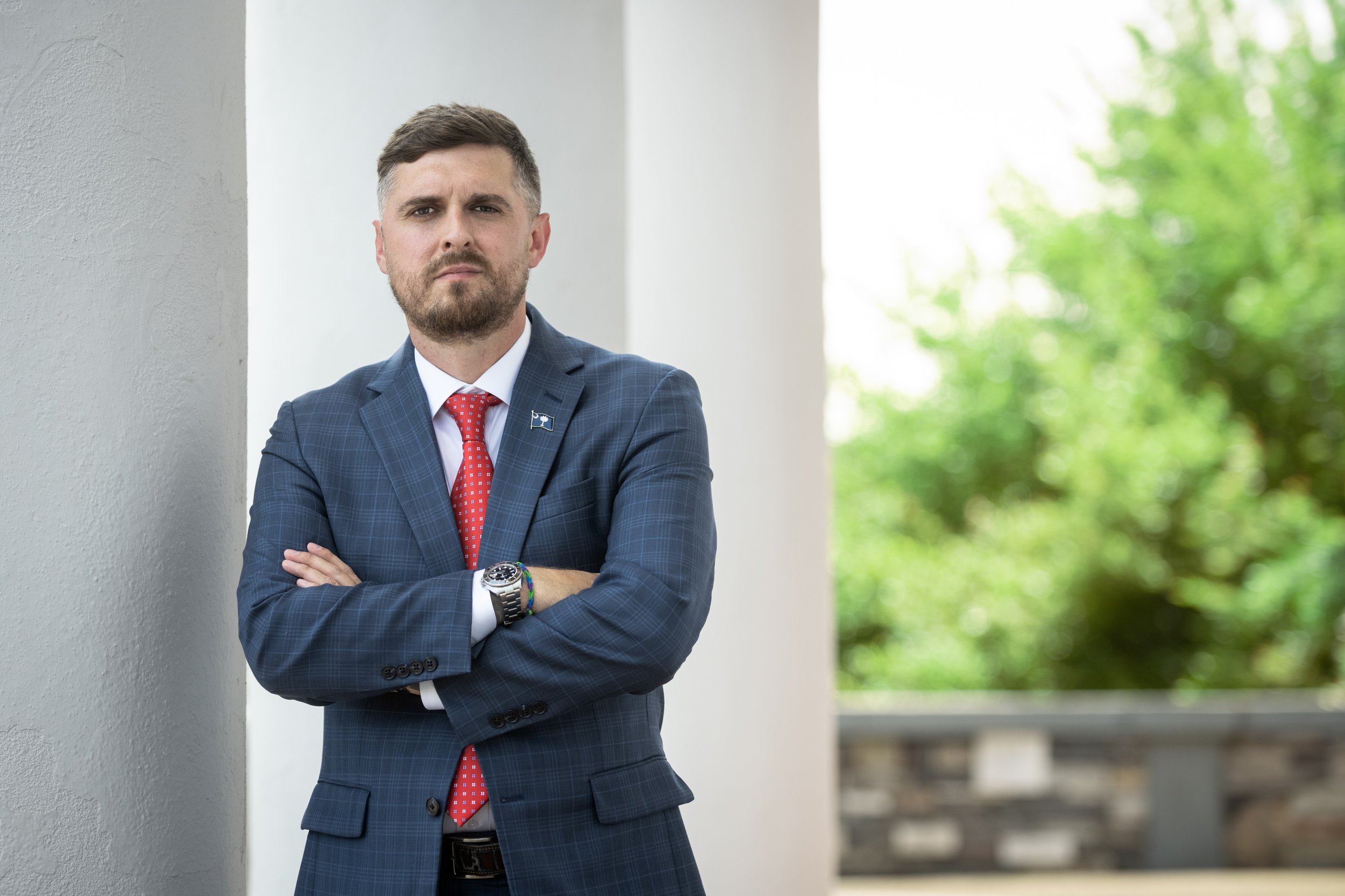A man in a blue checked suit with a red tie, standing with arms crossed, leaning against a white wall outdoors, with green trees and a railing in the background.
