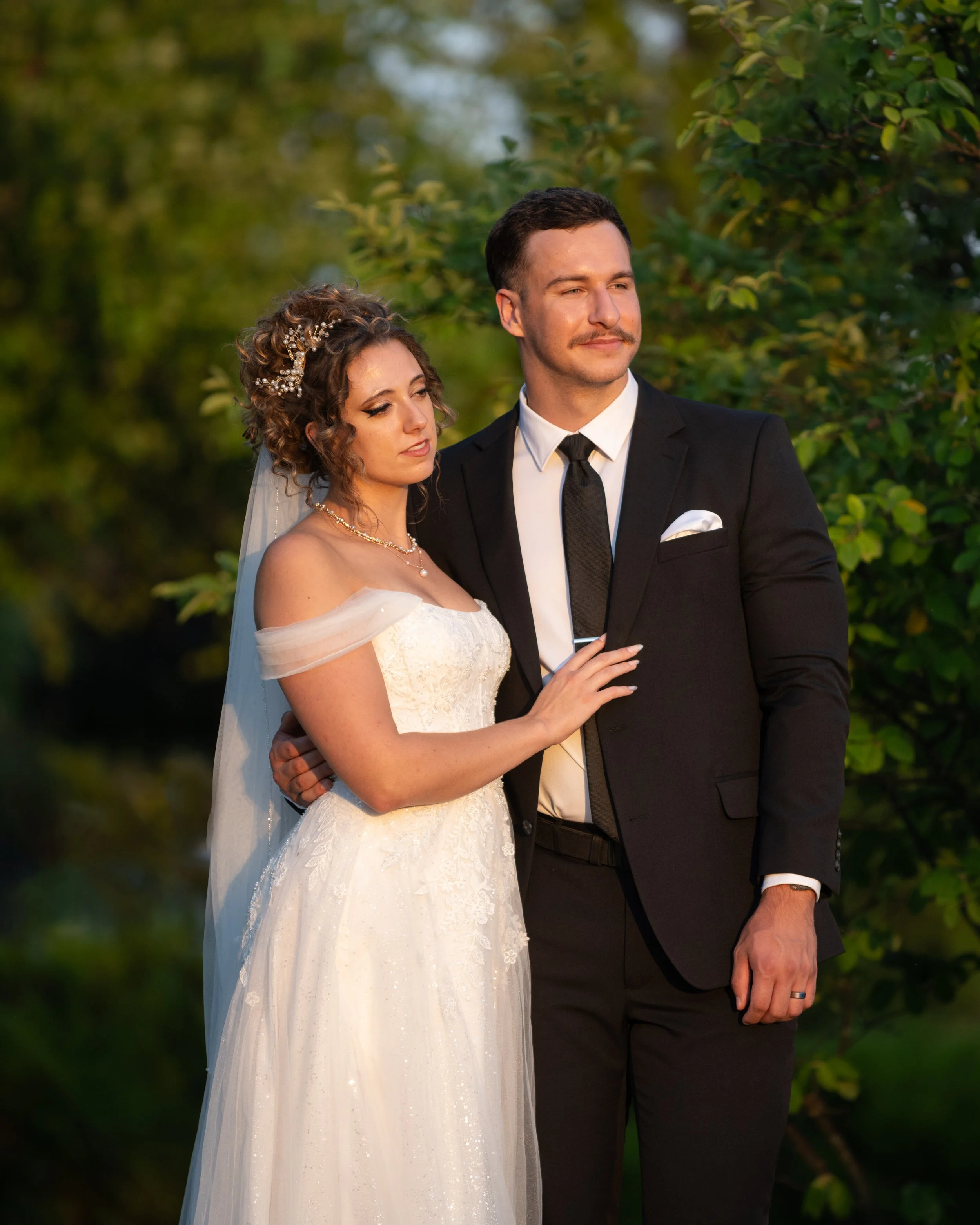 A bride and groom standing outdoors during sunset, with greenery in the background. The bride is in a white off-the-shoulder wedding dress with lace details, and the groom is in a black suit with a white shirt and a black tie.