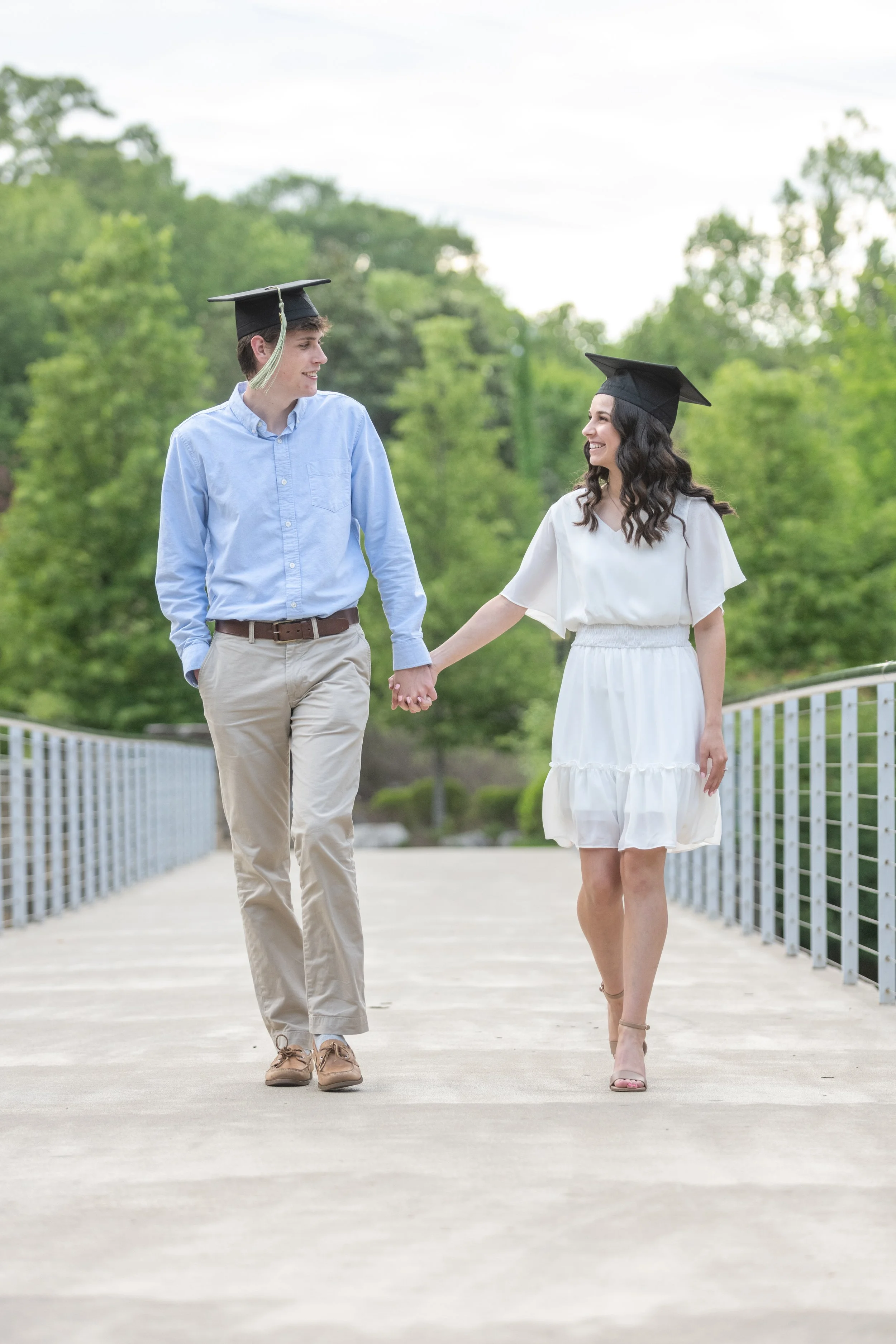 A young couple wearing graduation caps walking hand in hand on a bridge, smiling and looking at each other.
