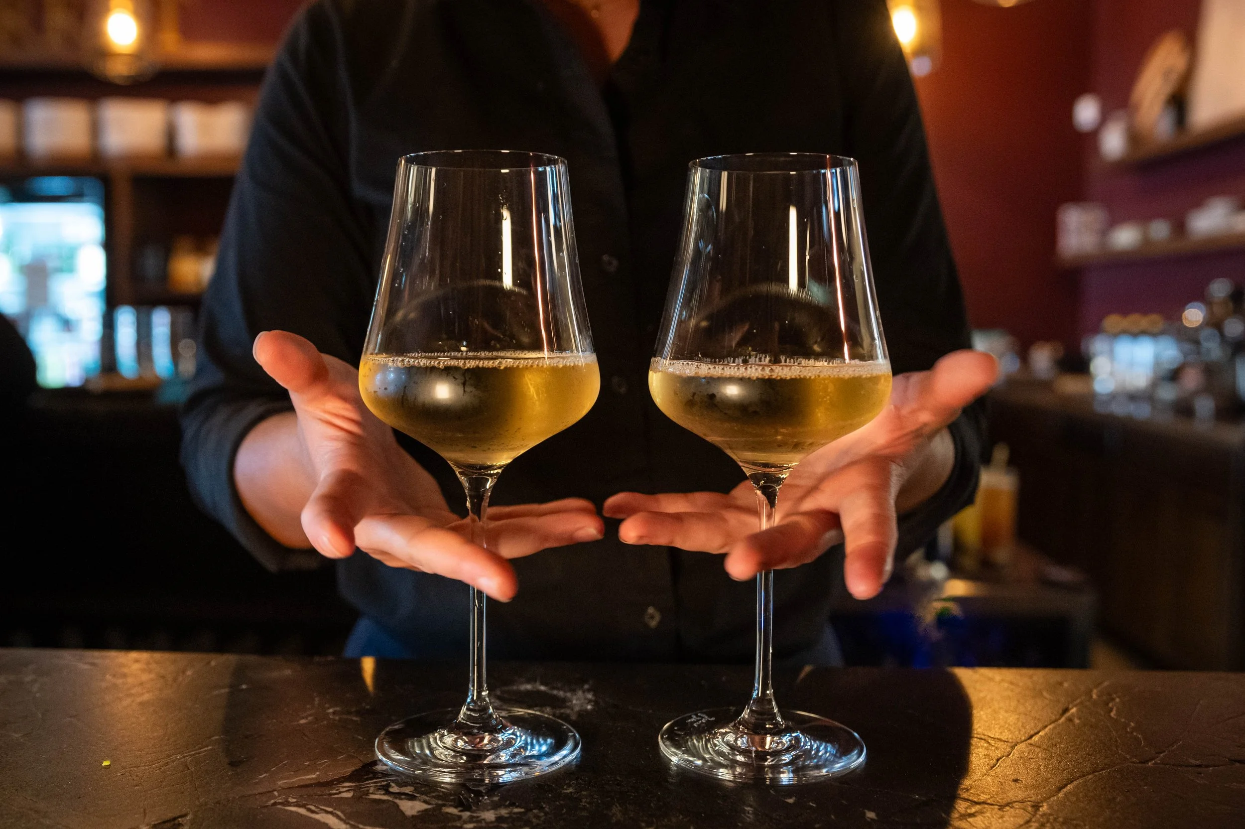 A person holding two glasses of white wine in a bar setting with warm lighting.