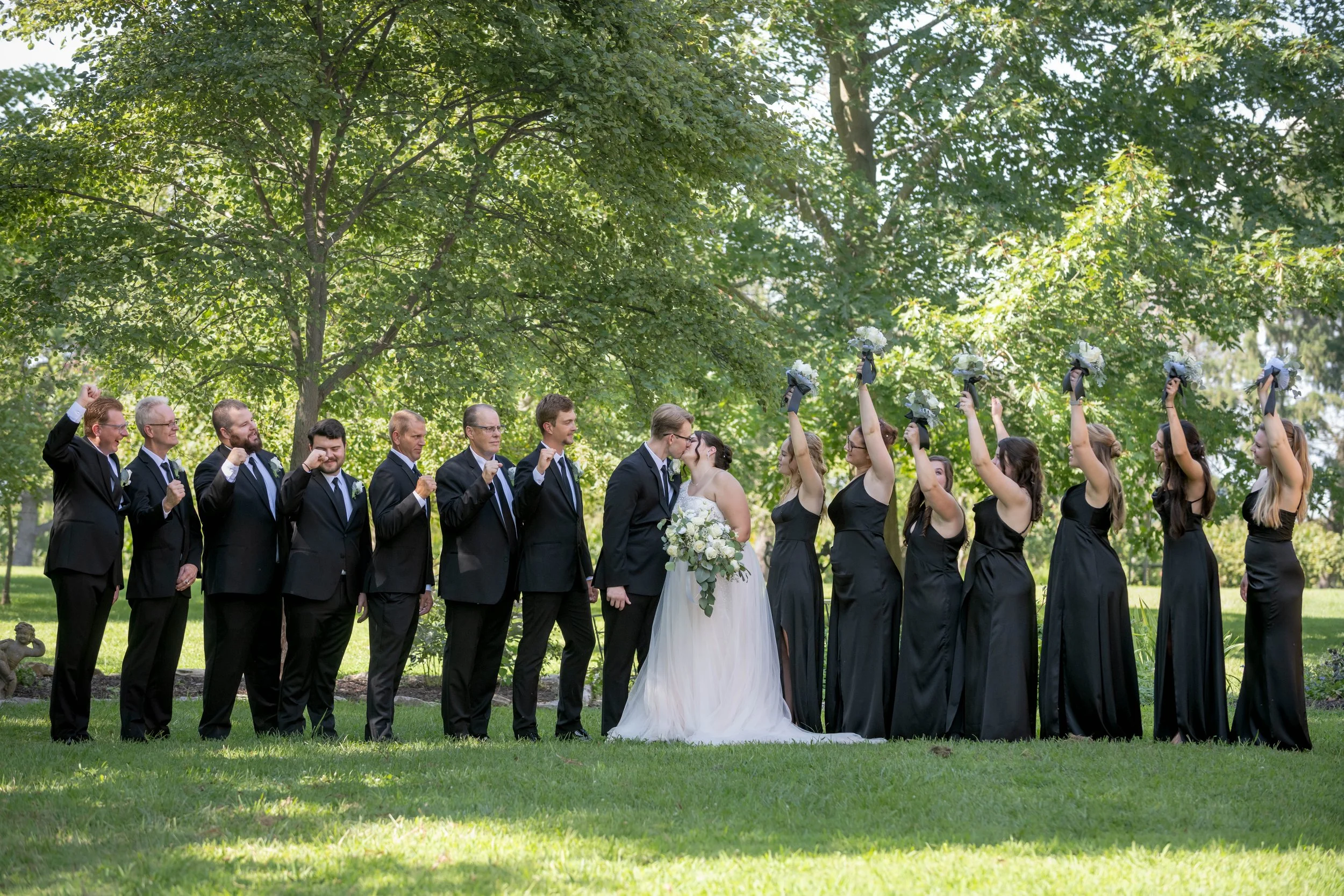 A wedding party standing outdoors under a large tree, with the bride and groom in the center kissing, surrounded by bridesmaids holding bouquets and groomsmen in black suits. The guests celebrate on a lush green lawn.