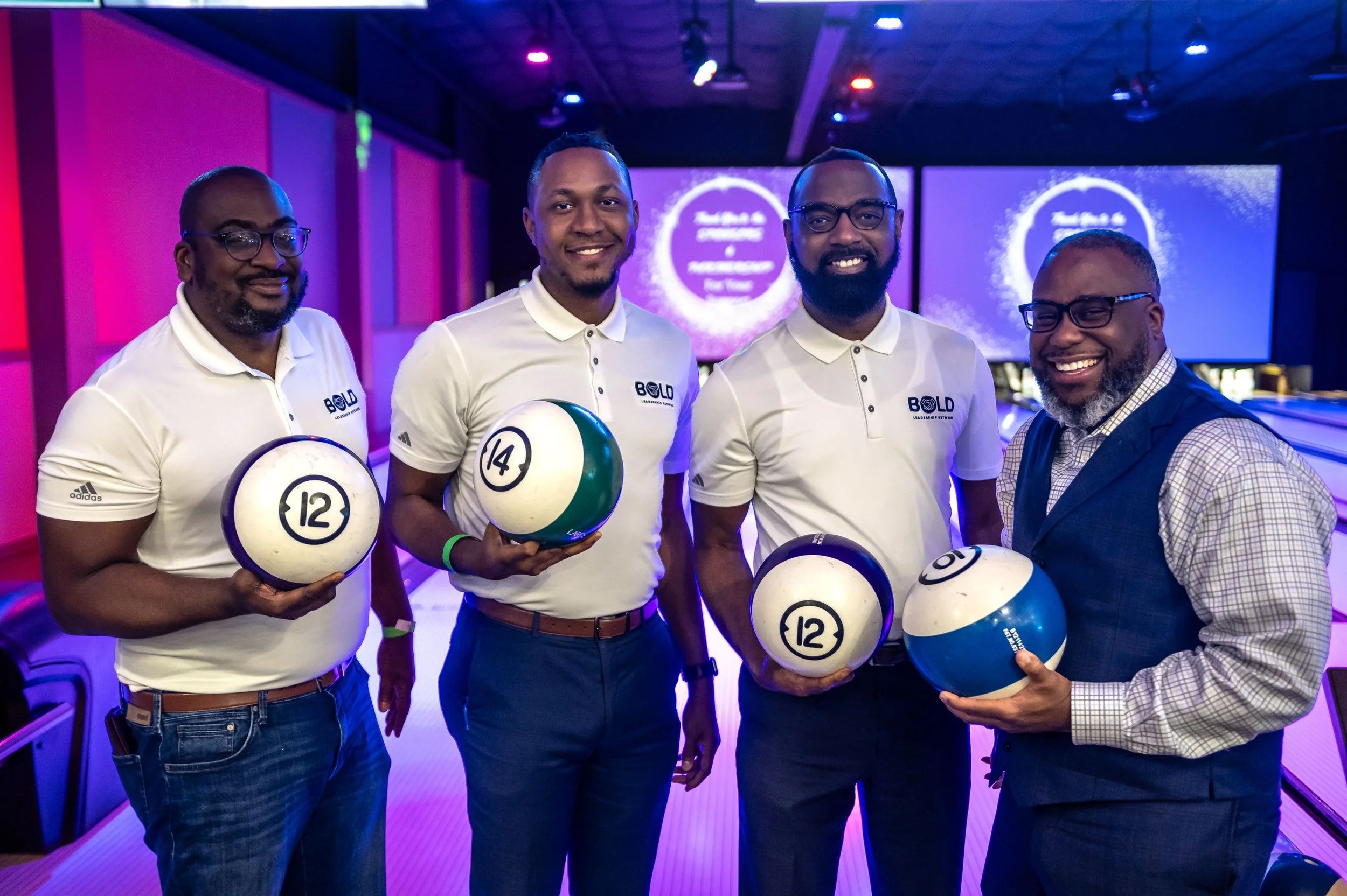 Four men at a bowling alley holding bowling balls, three of whom are wearing white polo shirts with a logo that says 'BOLD'. They are smiling and posing for the photo, with a background featuring colorful lighting and large screens.
