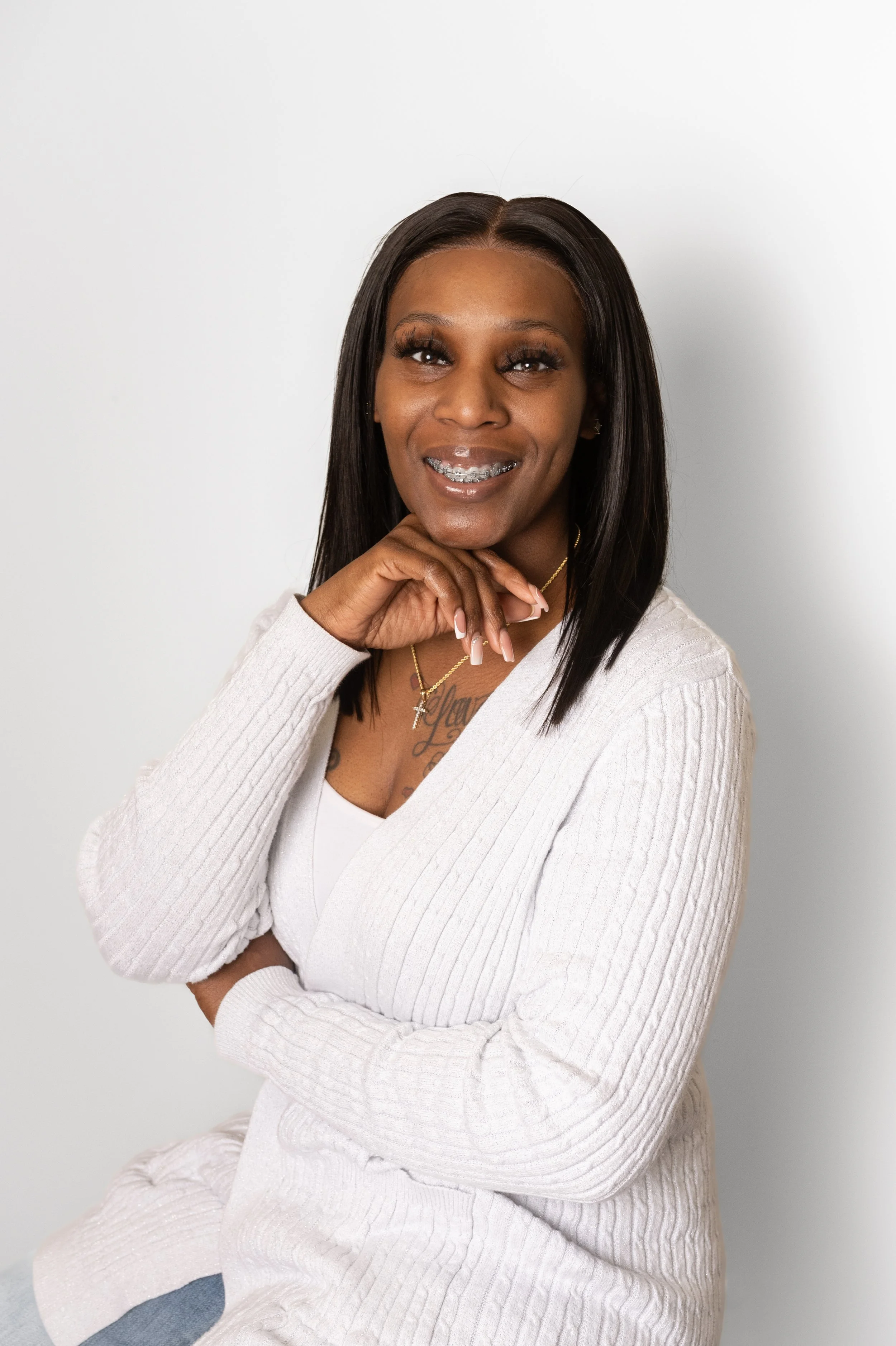 A woman with long straight black hair, wearing a white sweater, gold necklace, and braces, smiling and posing with her hand near her chin against a plain white background.