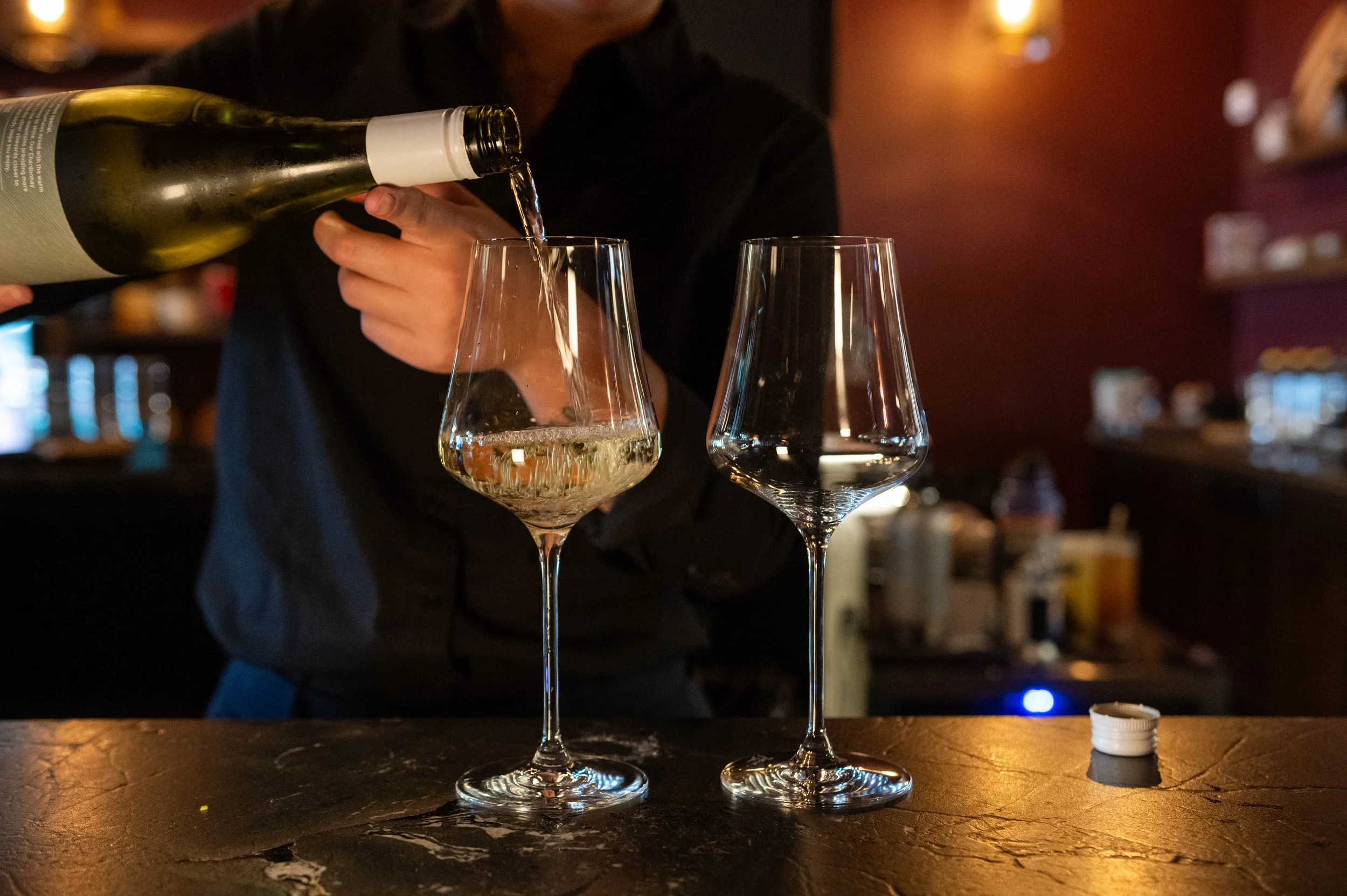 Person pouring white wine into a glass at a bar with another empty wine glass nearby.
