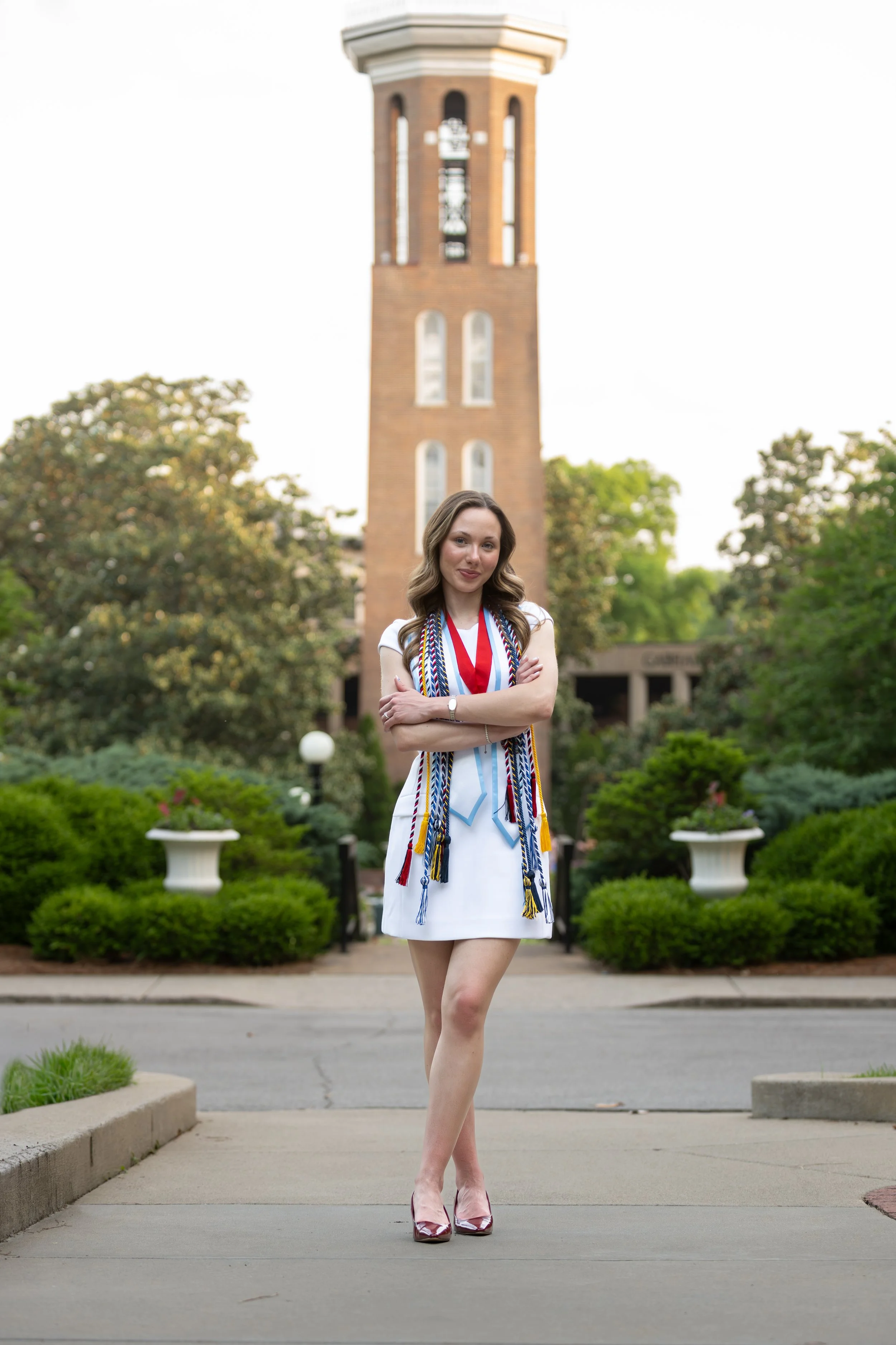 A young woman in a white dress standing outdoors in front of a campus clock tower, with greenery and potted plants around.