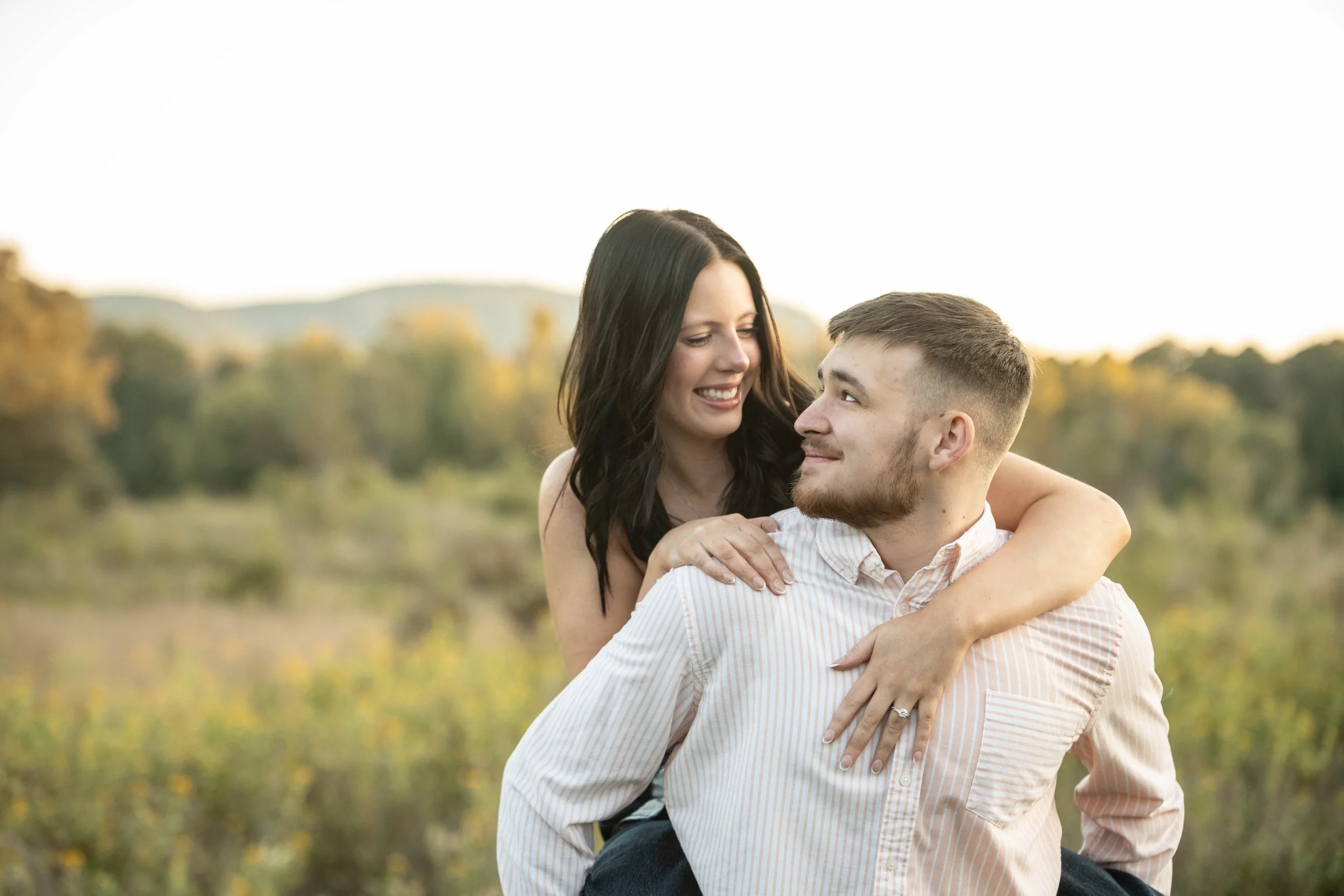 A woman with dark hair and a man with short hair and facial hair look at each other affectionately outdoors during sunset, with a blurred natural landscape in the background.