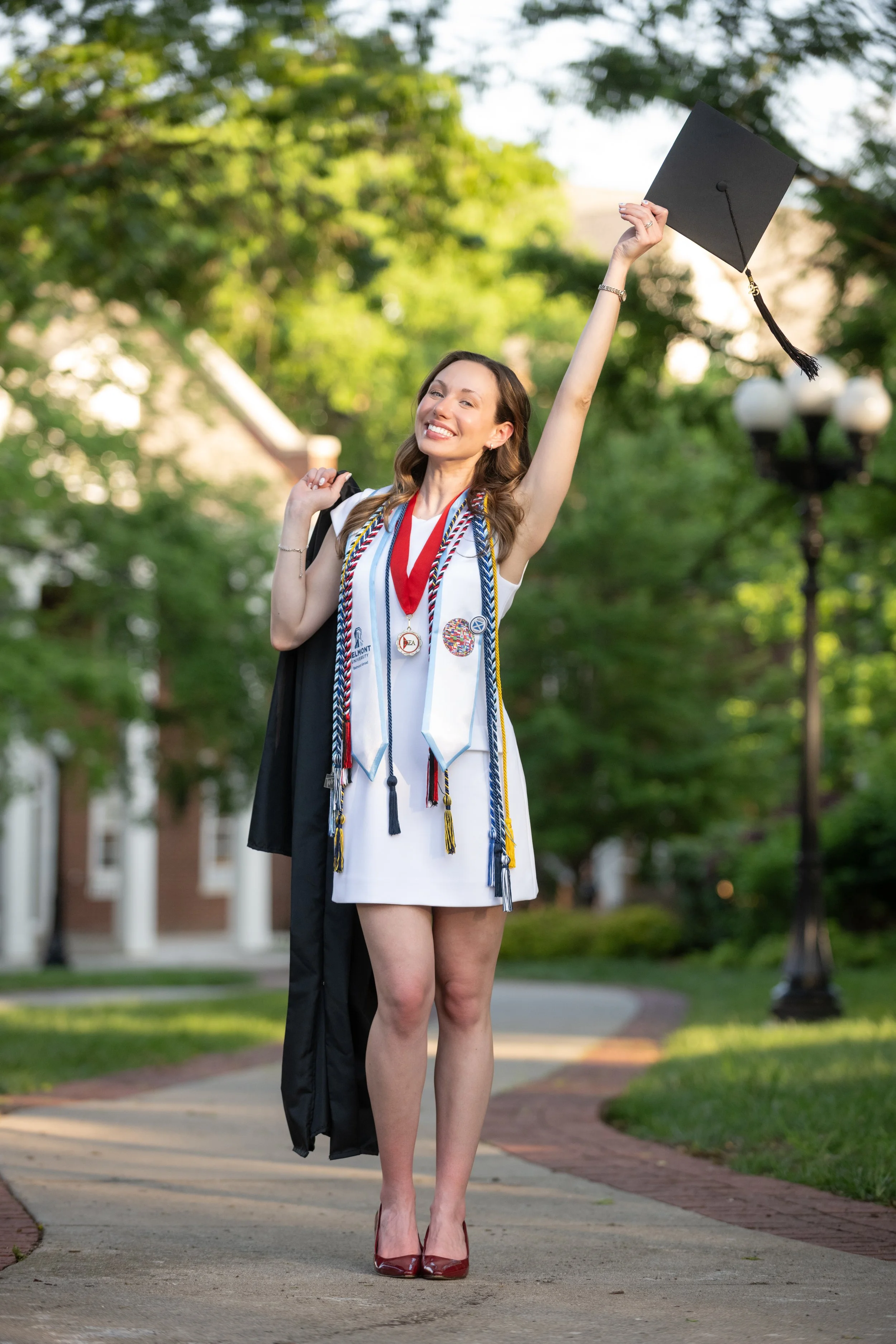 A young woman in graduation attire celebrating outdoors, wearing a white dress and multiple cords and medals, holding her cap high, smiling, with a campus pathway and trees in the background.