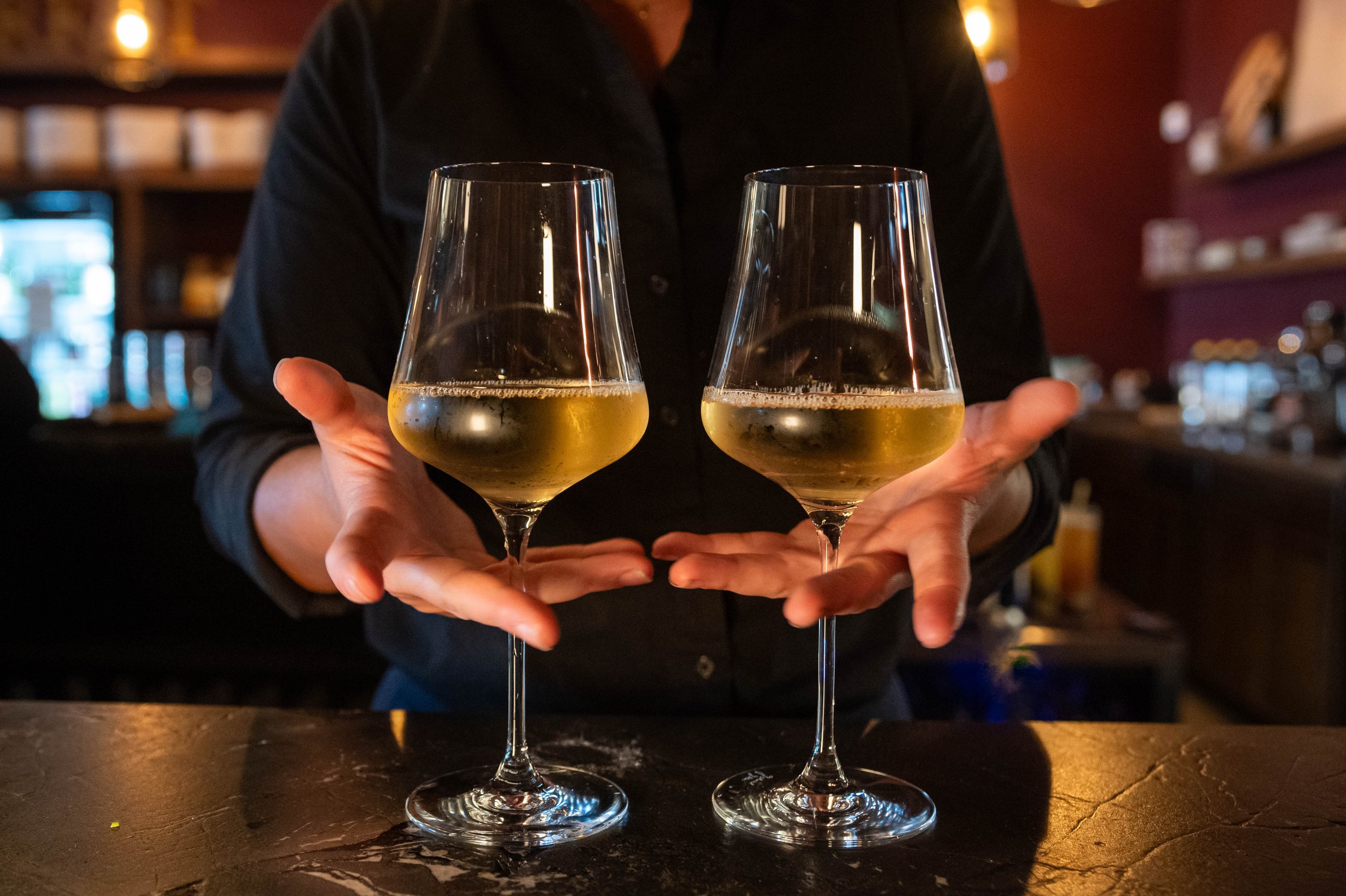Bartender holds out two tall wine glasses filled halfway with white wine.