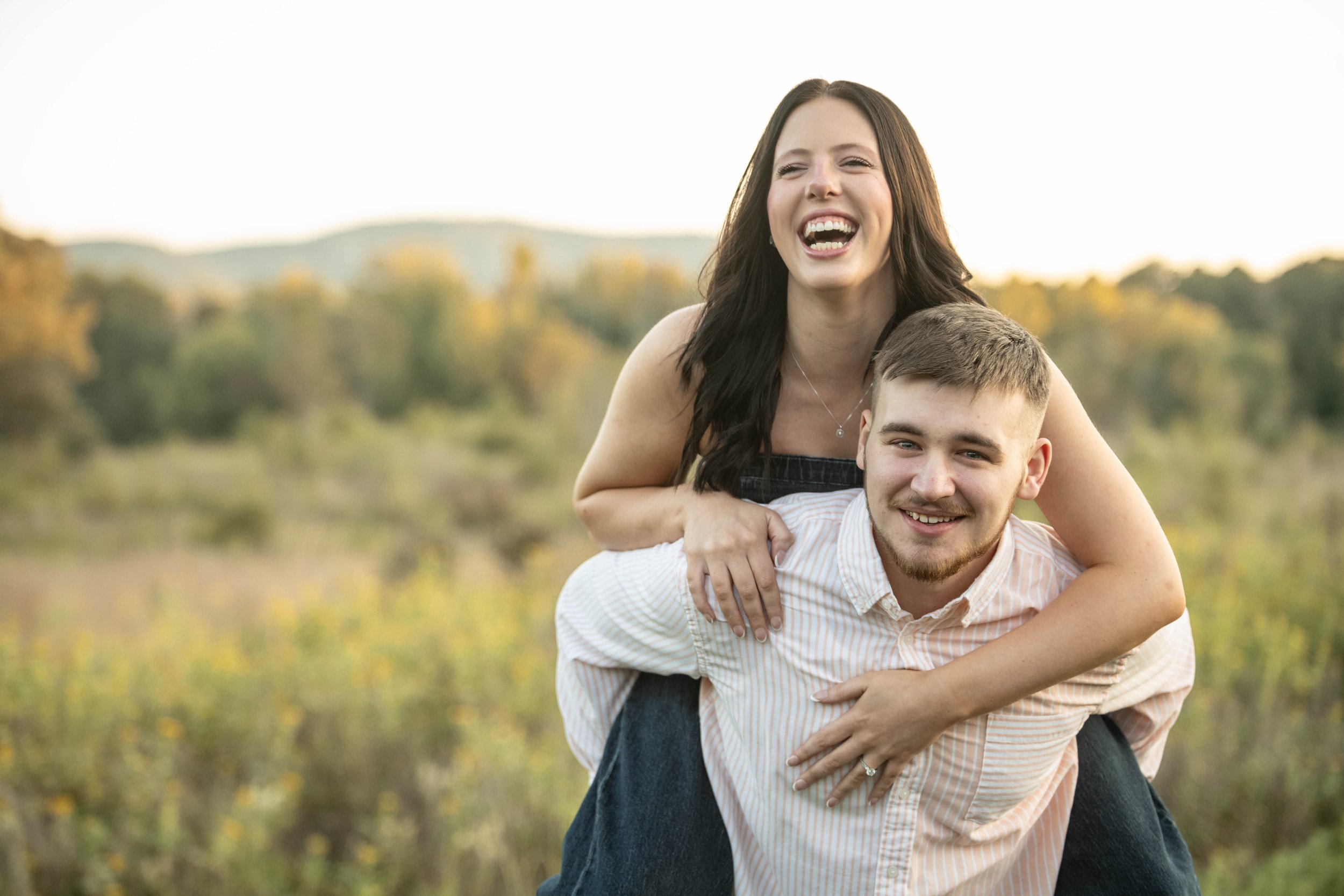 A woman giving a piggyback ride to a man in an outdoor natural setting with trees and mountains in the background, both smiling and enjoying the moment.