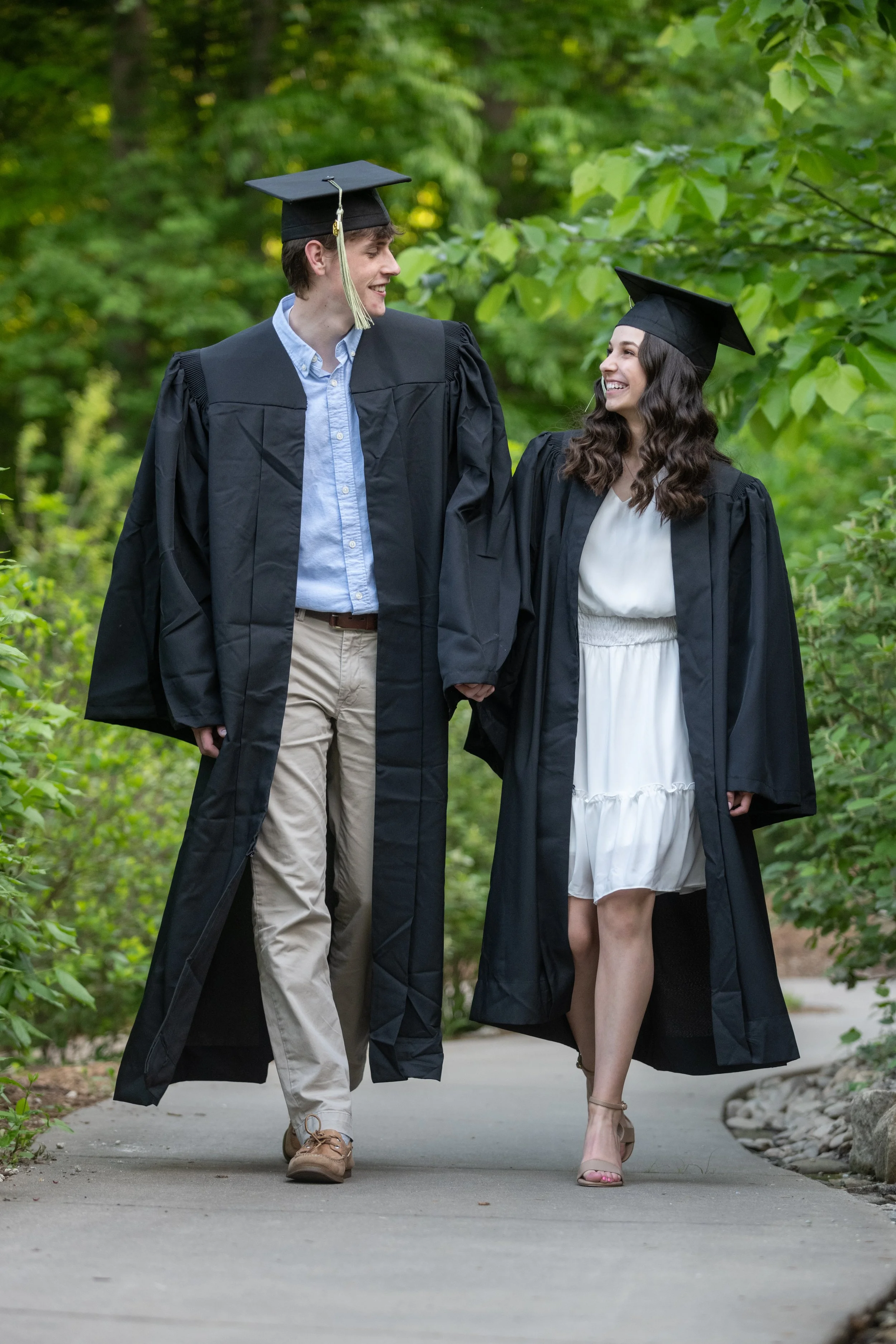 Two graduates, a man and a woman, holding hands and smiling while walking outdoors in graduation gowns and caps.