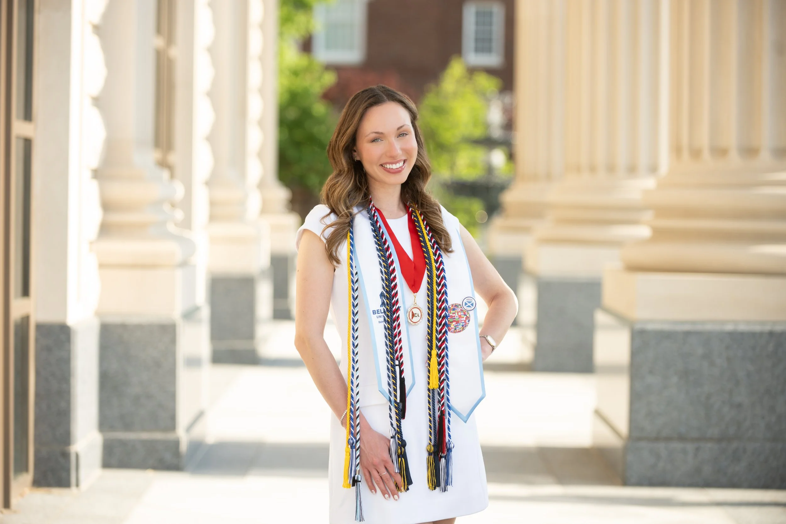 A young woman in a white dress standing outdoors, wearing various graduation cords and medals, smiling, with a background of classical columns and greenery.