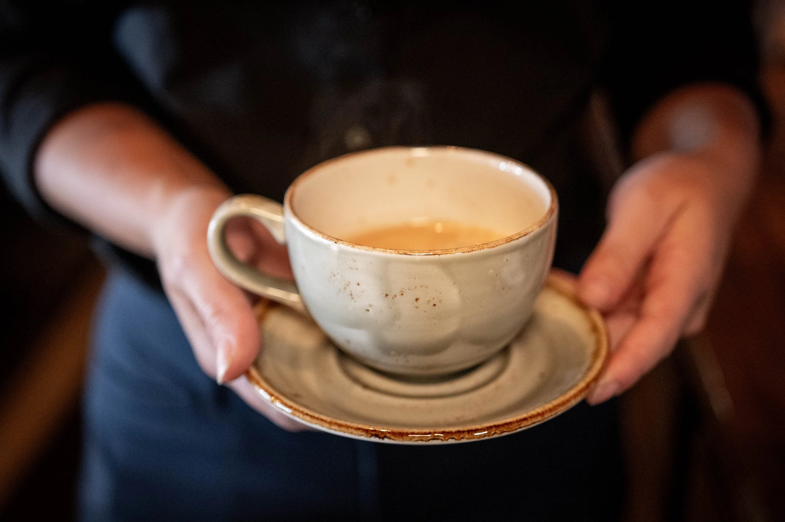 Person holding a beige ceramic cup with a saucer, filled with tea or coffee.