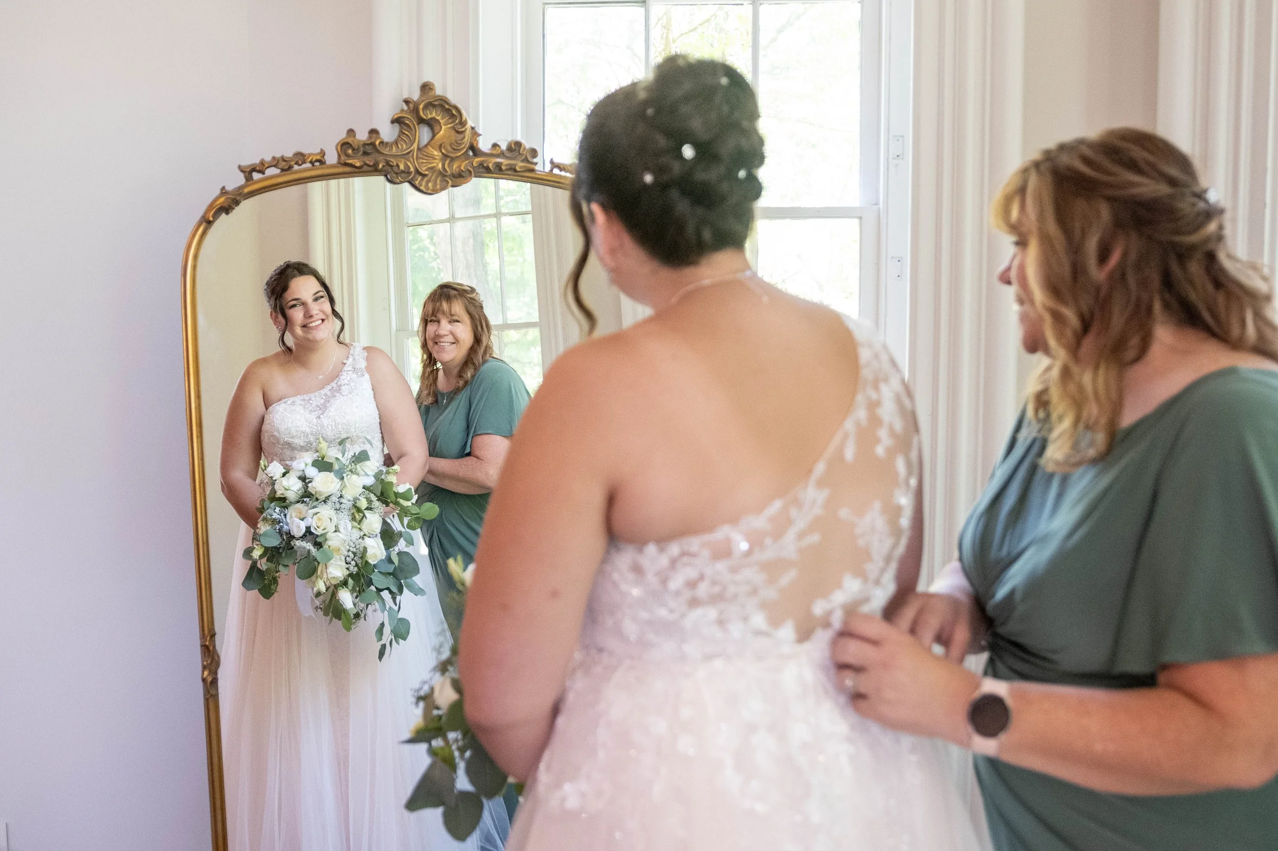 A bride in a white wedding gown holding a bouquet of white roses and greenery, smiling at her reflection in a large gold-framed mirror. Two women, possibly her mother and bridesmaid, stand nearby in a softly lit room with large windows, watching her 