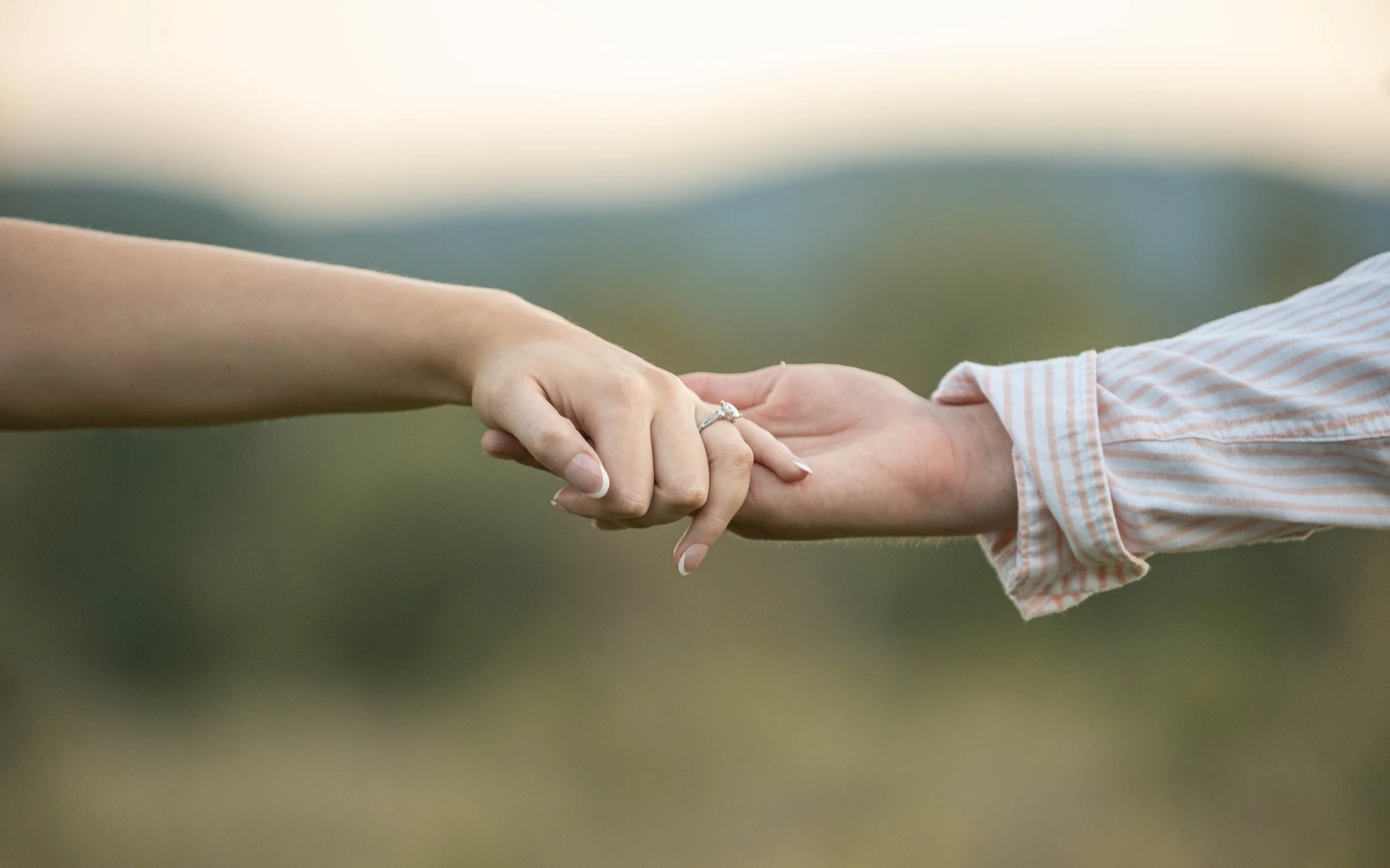 Close-up of a man and woman's hands holding each other, the woman wearing an engagement ring, outdoors with blurred green and brown background.