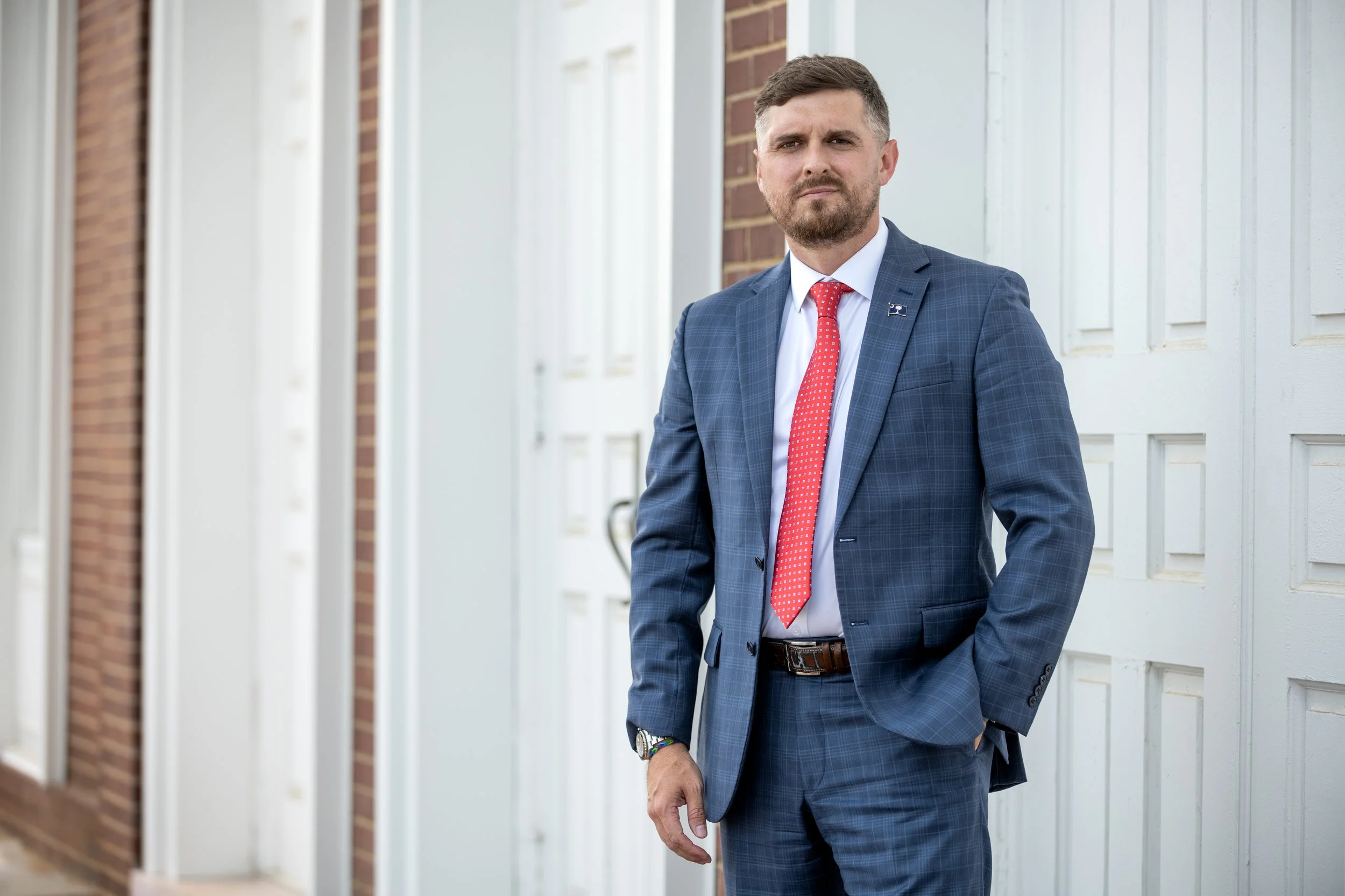 Man in a checkered blue suit with red tie standing outdoors in front of a white garage door and brick wall