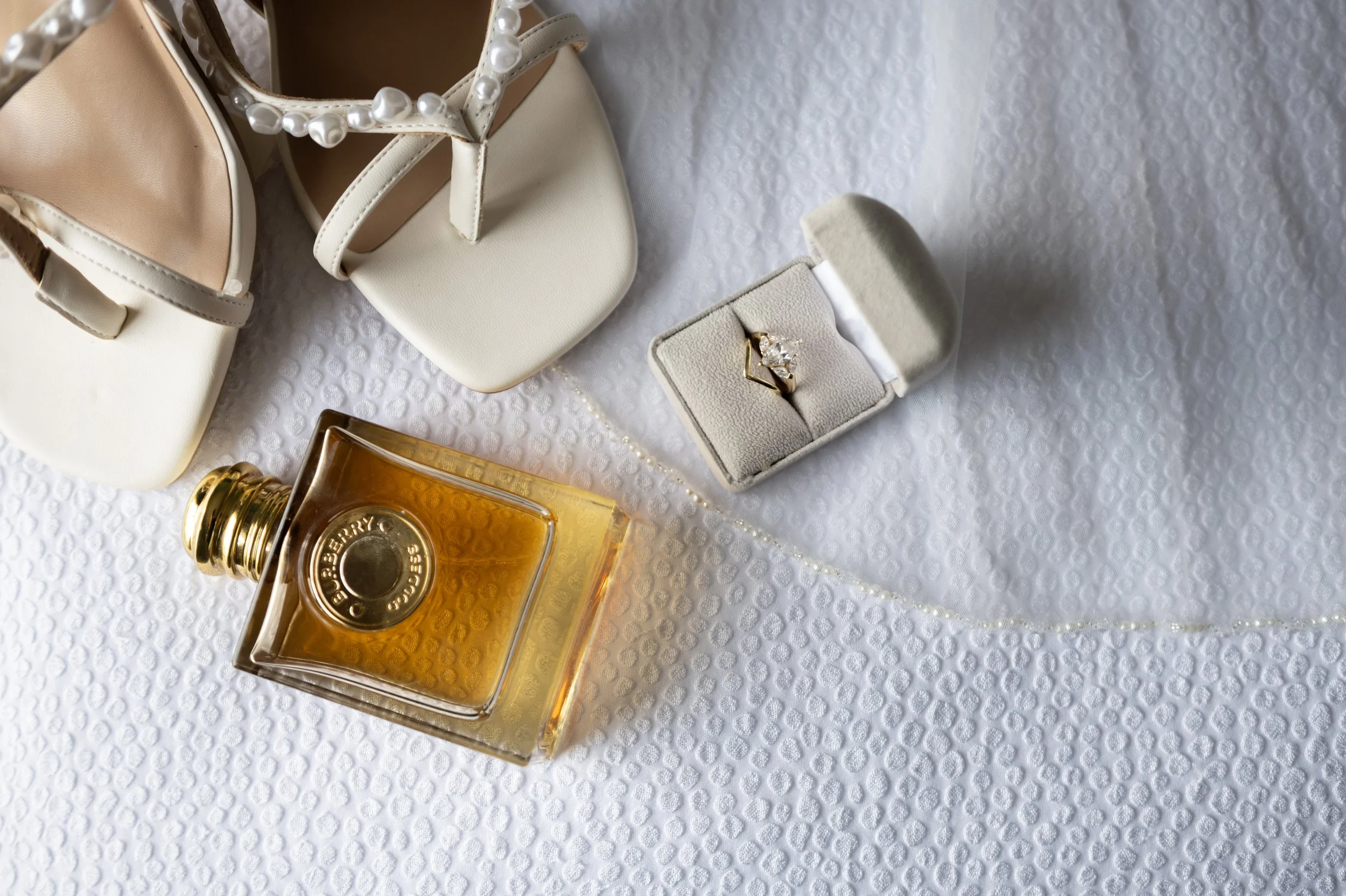 A pair of beige high-heeled shoes, a pearl necklace, a bottle of Burberry perfume, and a ring in a white jewelry box are arranged on a textured white surface.