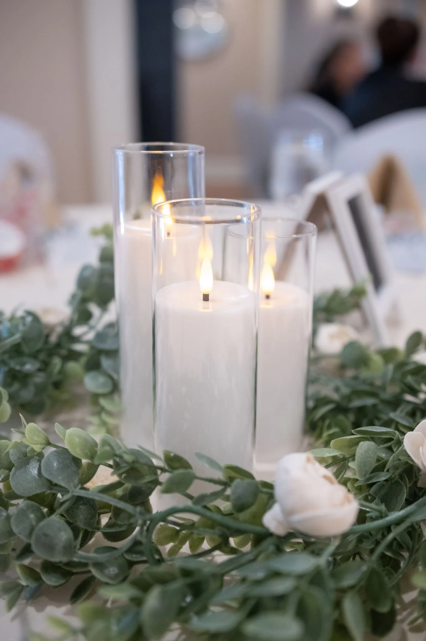 Close-up photo of three tall glass candles with flickering flames, surrounded by green foliage and a white flower, at a formal event or dinner setting.