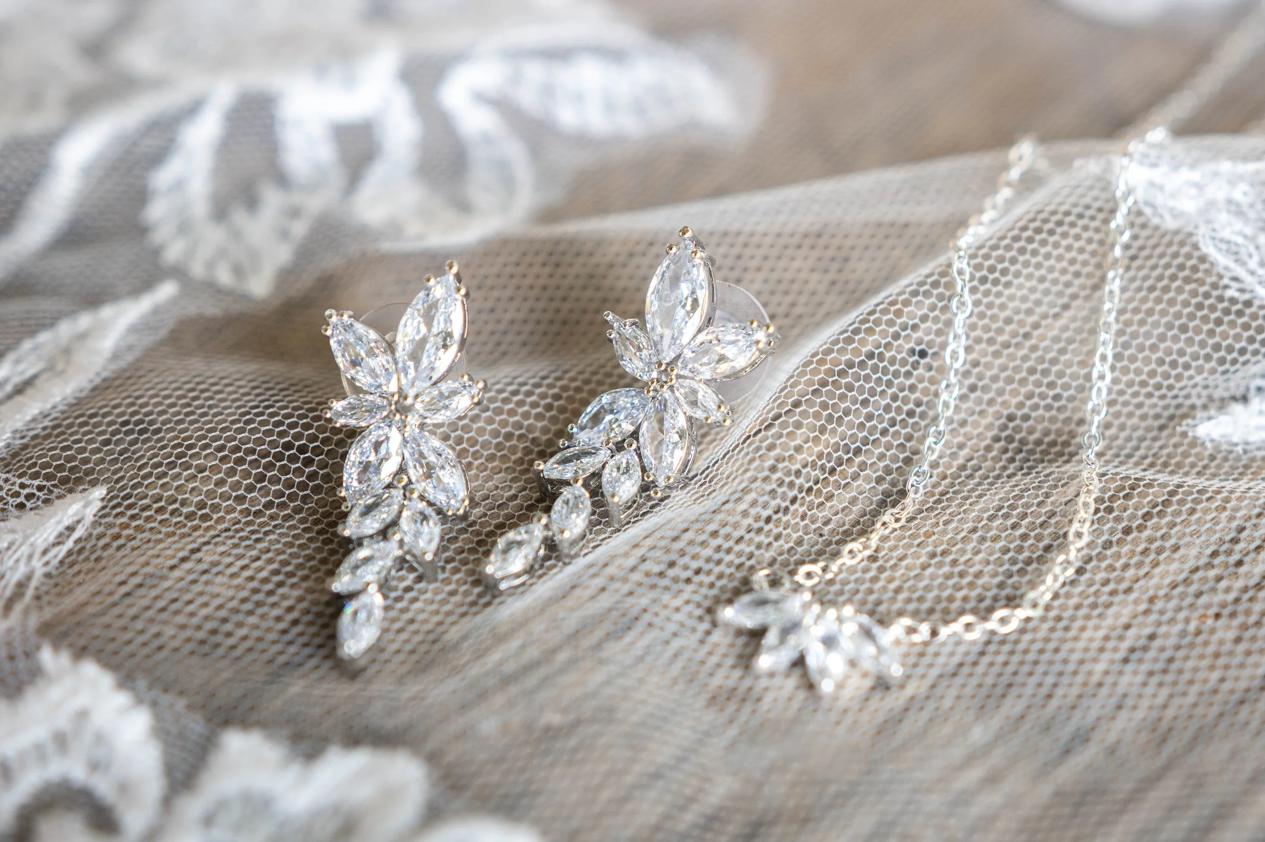 A set of silver jewelry with large diamond-like stones, including a pair of earrings and a necklace, displayed on a lace fabric background.