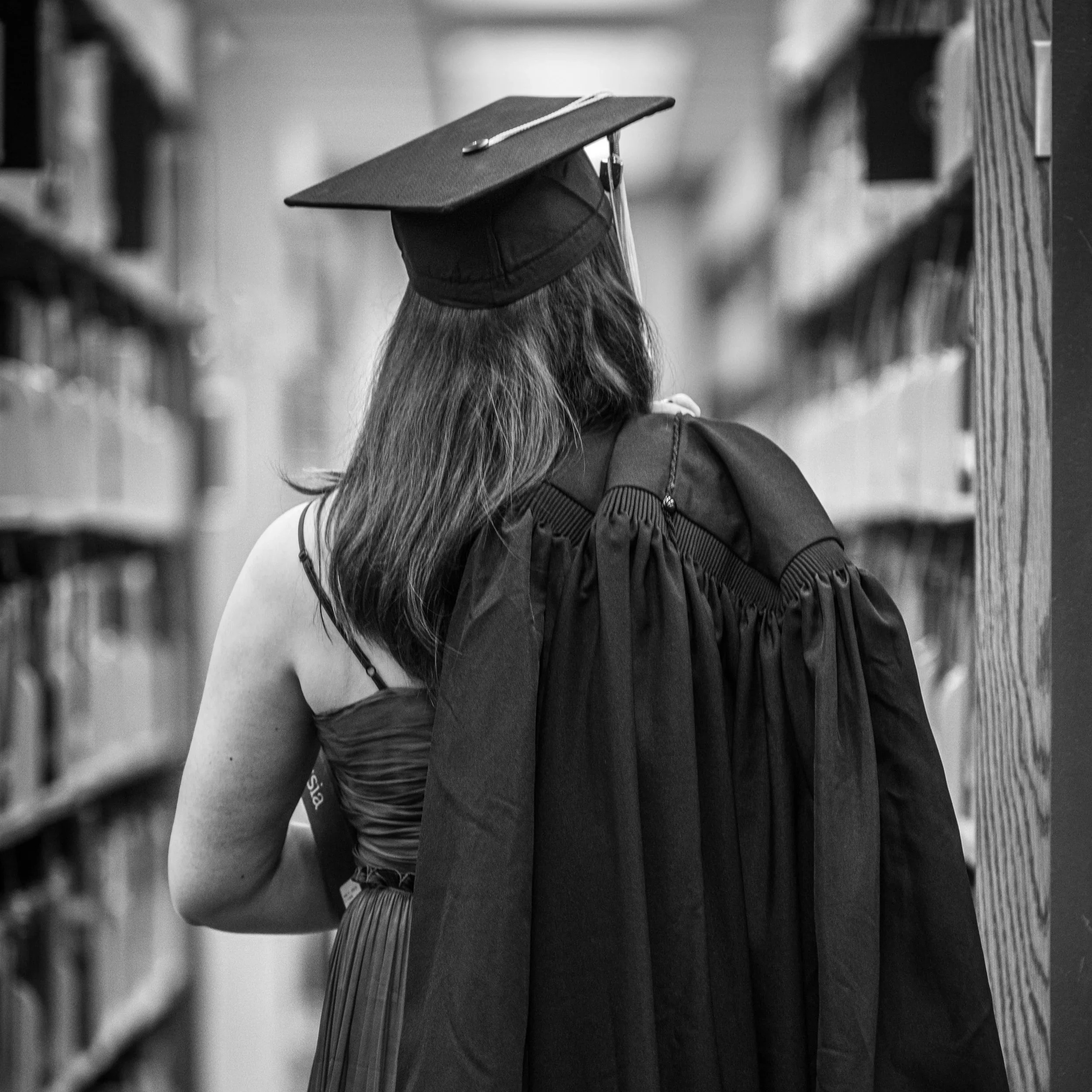A woman wearing a graduation cap and gown in a library aisle, looking at books on the shelves.