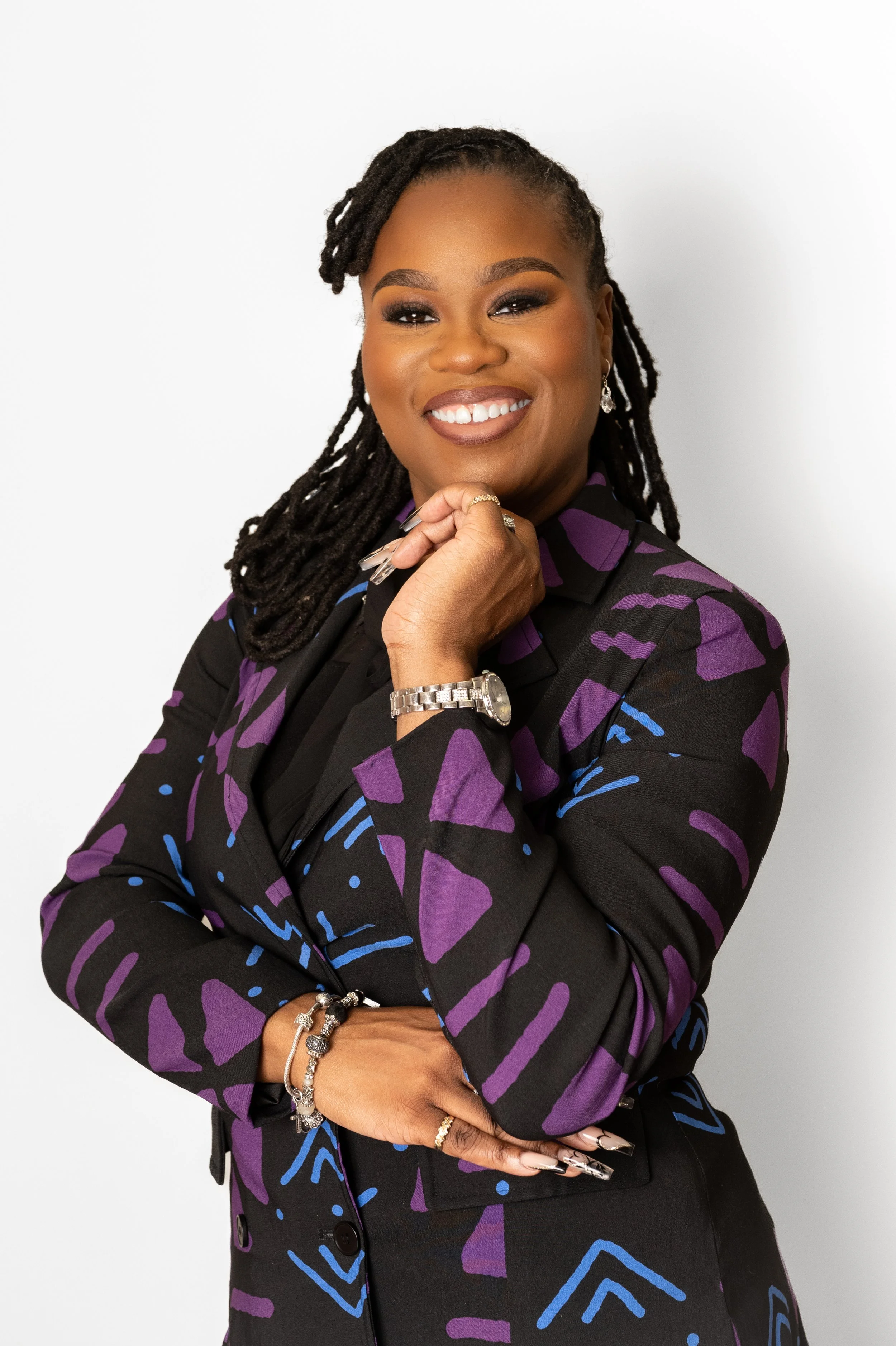 A smiling woman with long dreadlocks, wearing a black blazer with purple and blue abstract patterns, silver jewelry, and a wristwatch, standing against a plain white background.