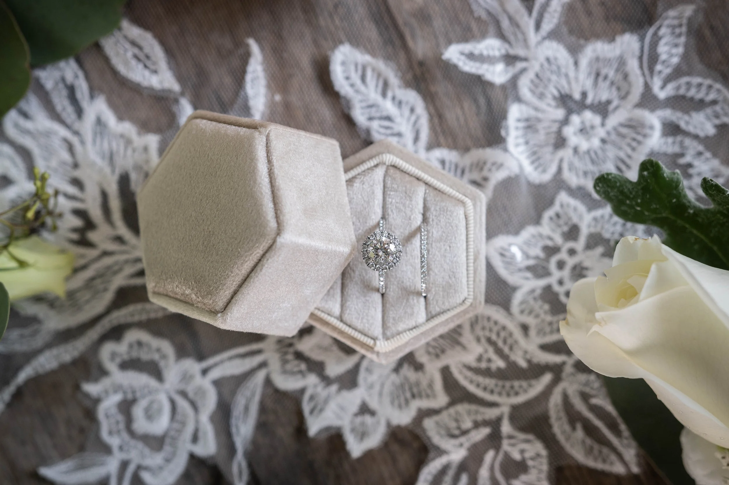 A diamond engagement ring in a velvet ring box, with a matching wedding band, on a lace tablecloth with flowers nearby.