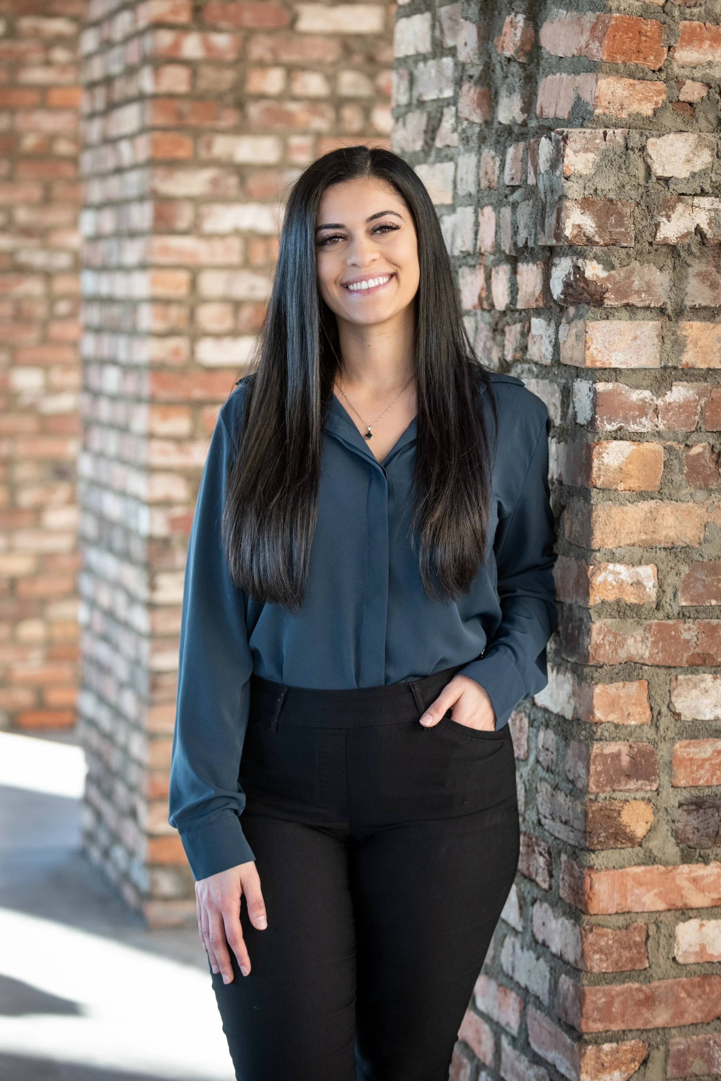 A smiling young woman with long dark hair, wearing a navy blue blouse and black pants, standing against a brick column outdoors.