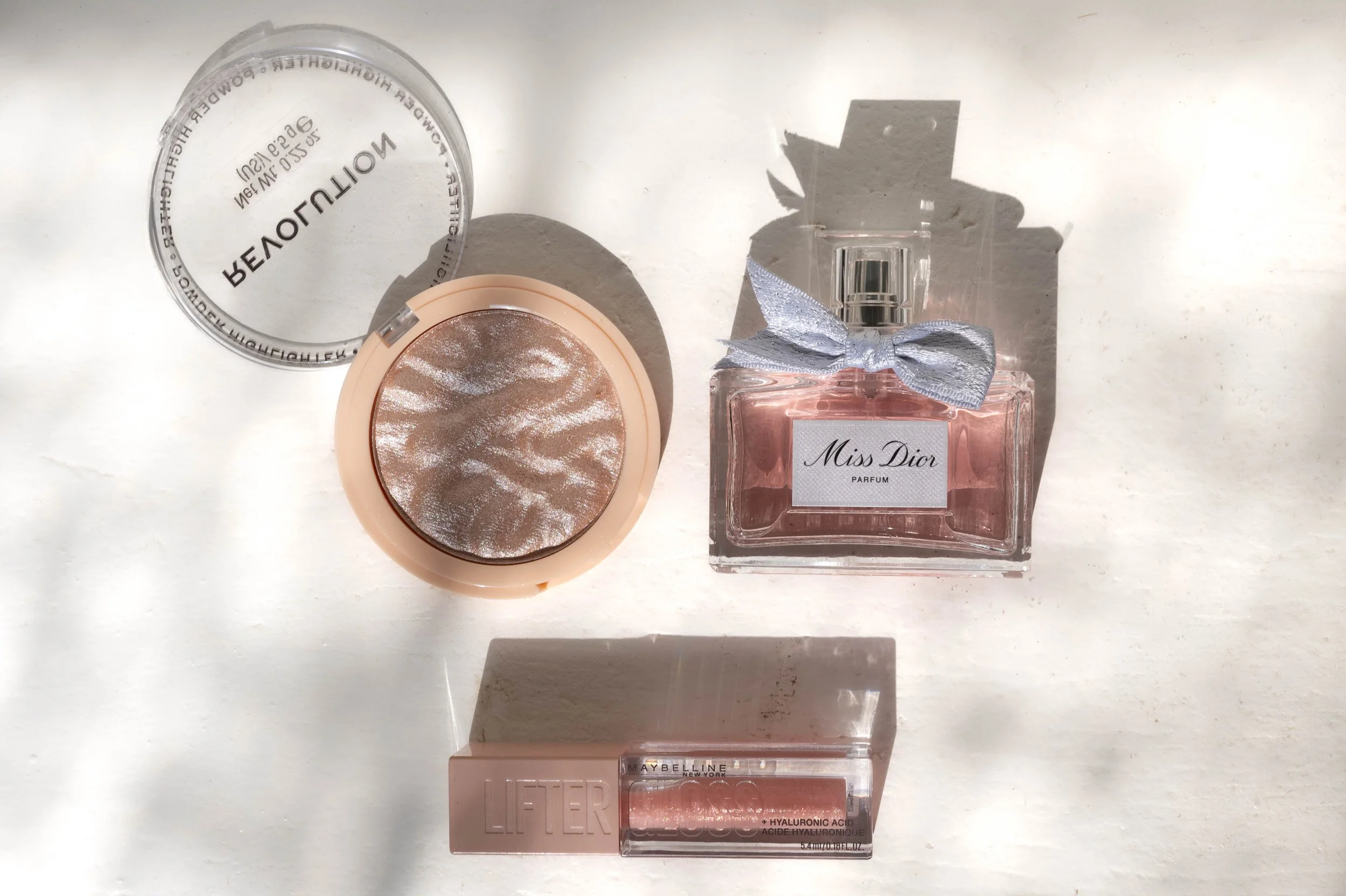 Collection of makeup and perfume items on a white surface, including a compact mirror with a shiny, circular metallic makeup product, a bottle of Miss Dior perfume with a silver bow, and a Maybelline lipstick in a transparent casing.