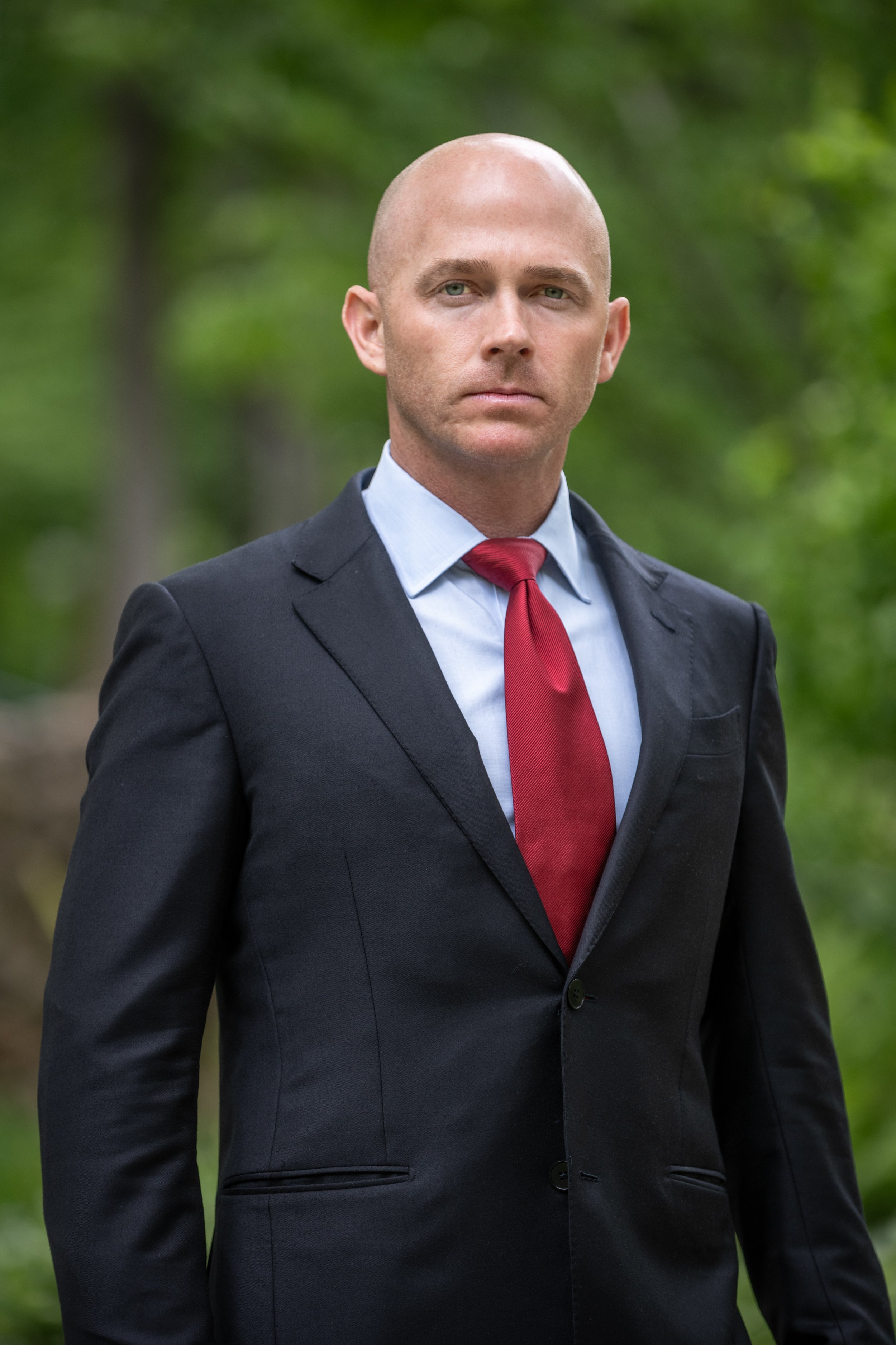 A bald man in a black suit, light blue shirt, and red tie standing outdoors with green blurred trees in the background.