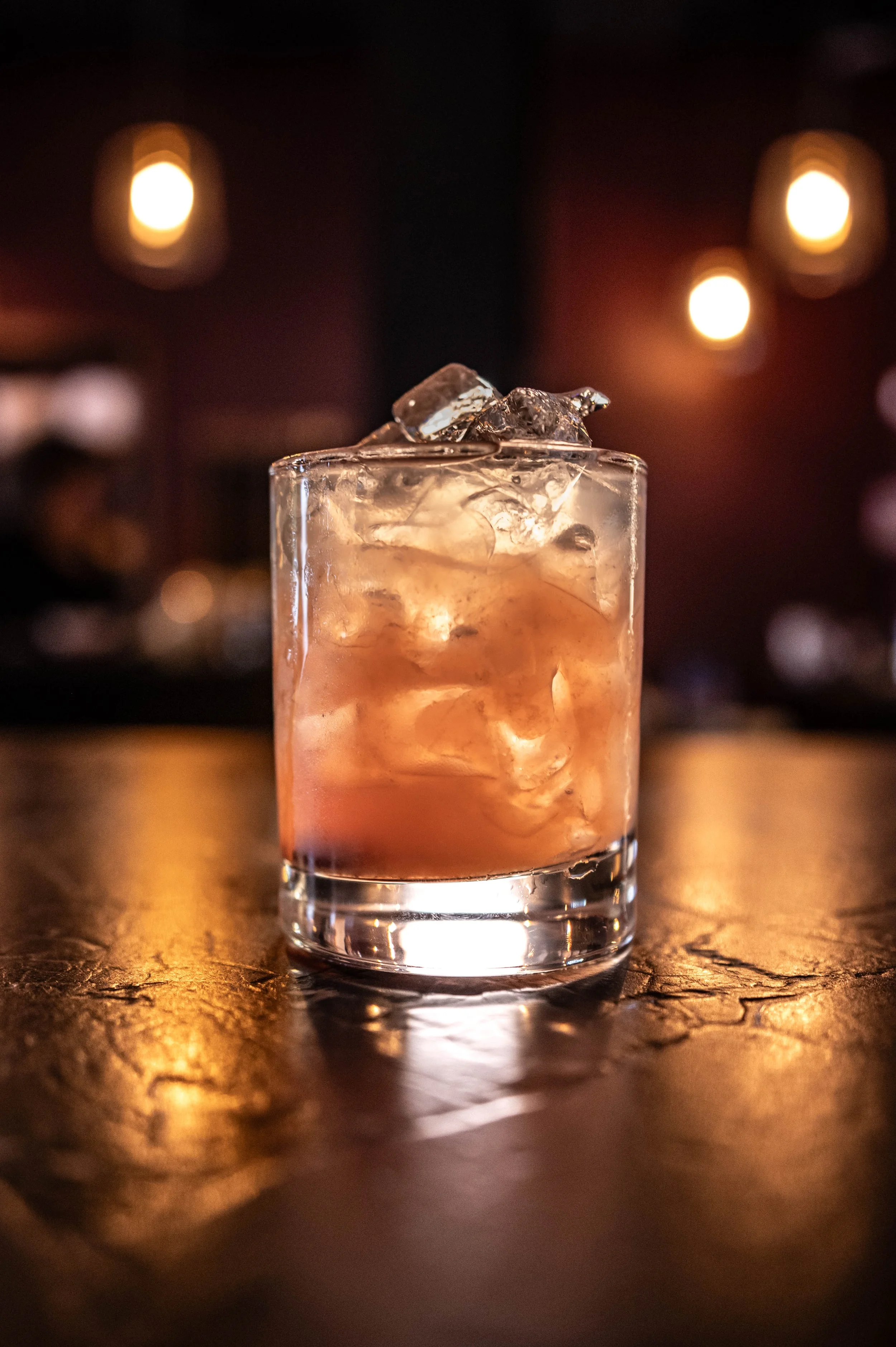 A glass of cocktail with ice cubes on a dark wooden bar surface, illuminated by warm ambient lighting in the background.