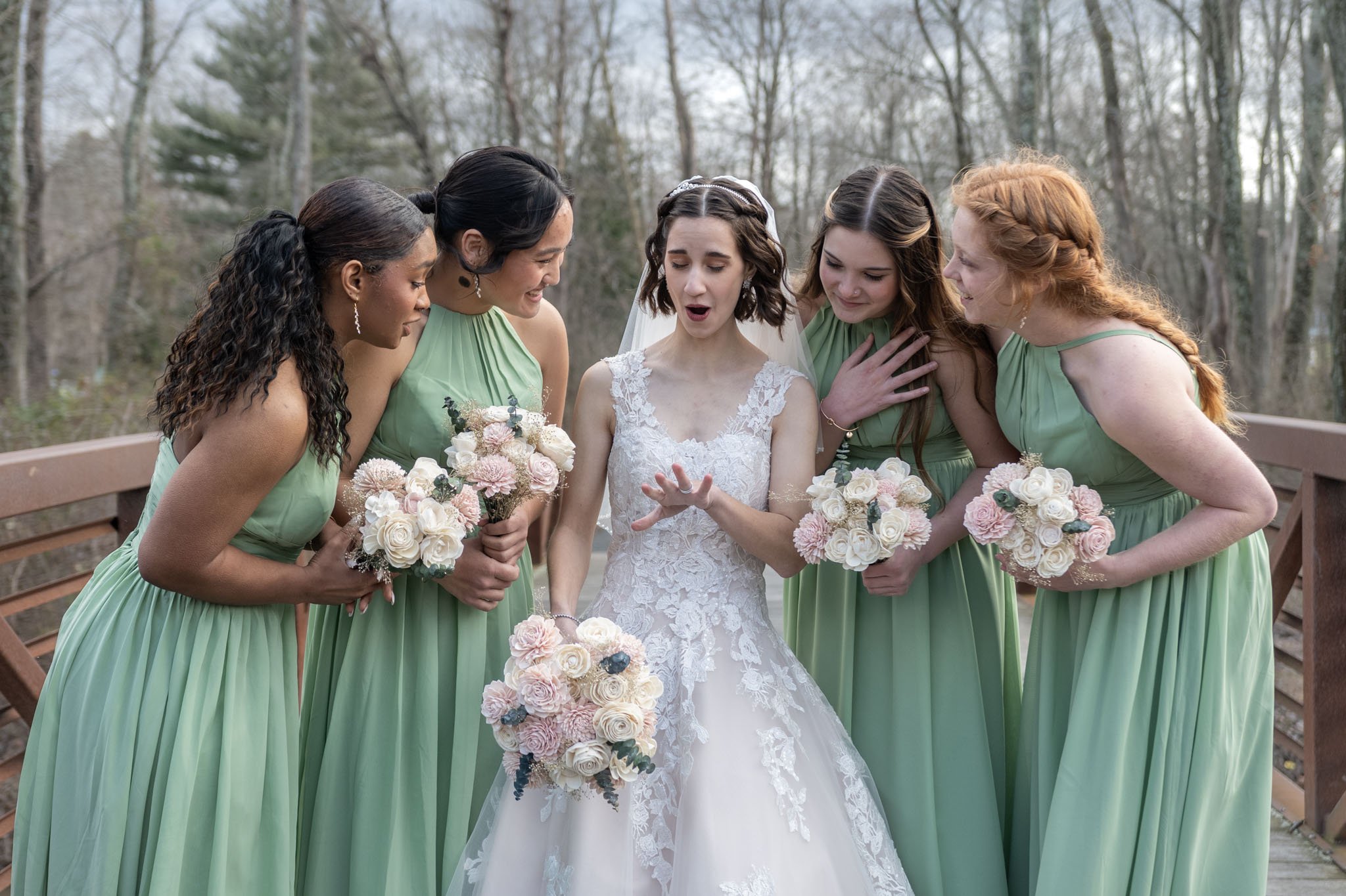 Bridal party with five women, four in matching green dresses, gathered around the bride on a wooden bridge outdoors, with trees in the background. The bride wears a white lace wedding gown and hold a bouquet, while the other women hold bouquets of fl